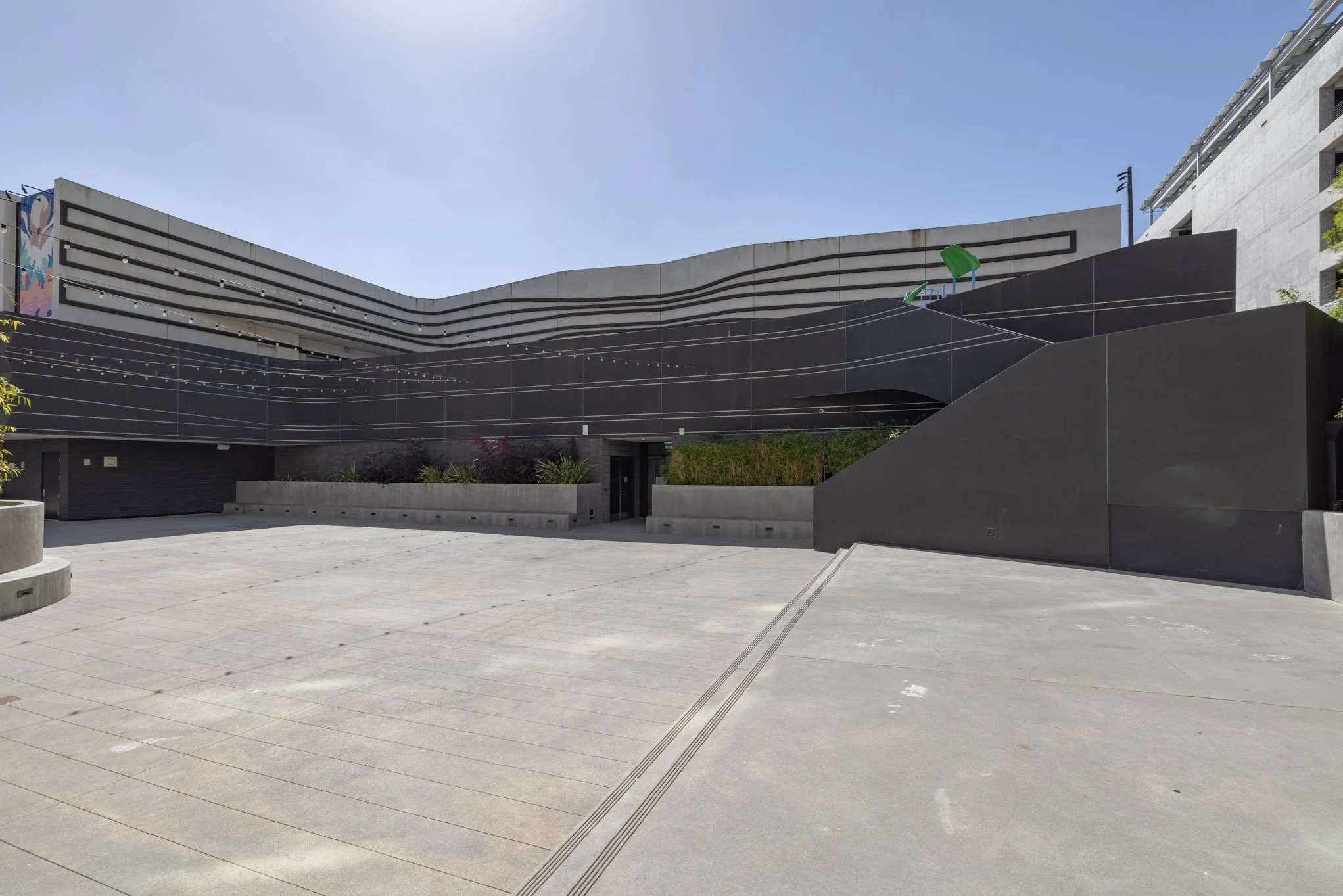 Modern building with dark exterior and abstract lighter lines, planter boxes, and stairs leading up to the entrance, under a blue sky.