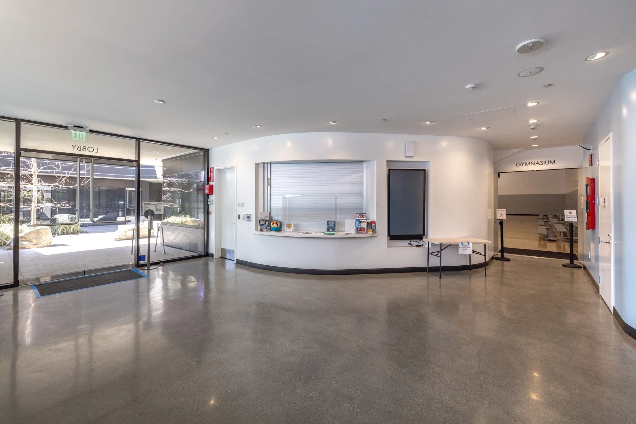 Interior of a school lobby with glass doors, a reception window with brochures, and a hallway leading to a gymnasium.