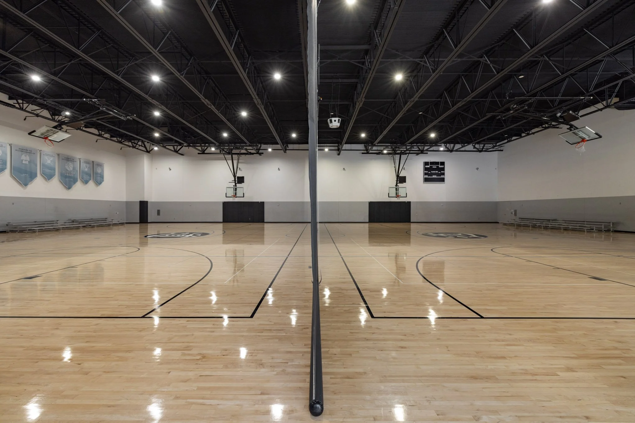 Empty indoor basketball court with a net dividing the court in half, polished wood floors, and a high ceiling with bright overhead lights.
