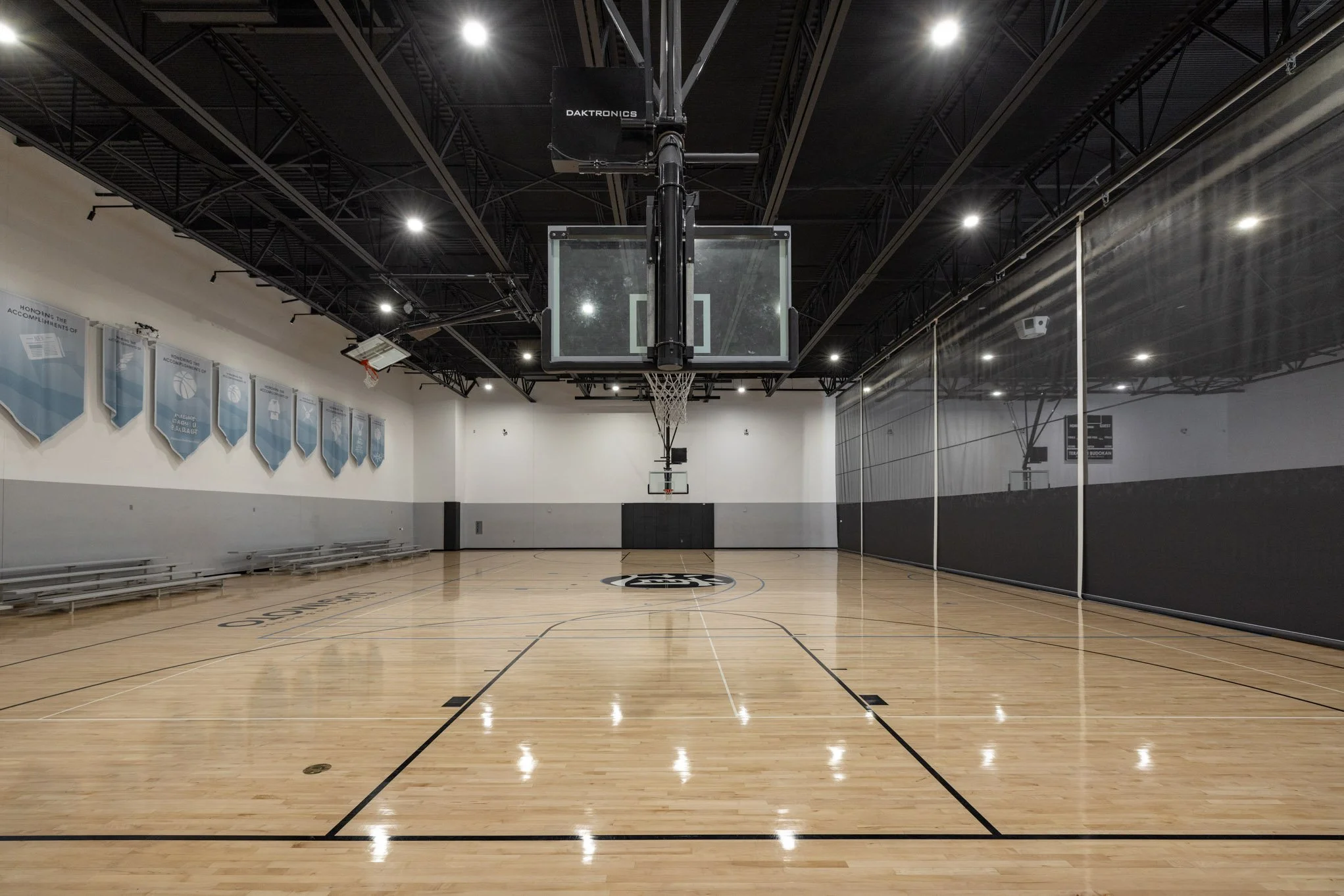 Empty indoor basketball court with polished wooden floor, basketball hoops, and a black padded backboard in the center. The court is well-lit with overhead lighting and has bleachers on the left side.