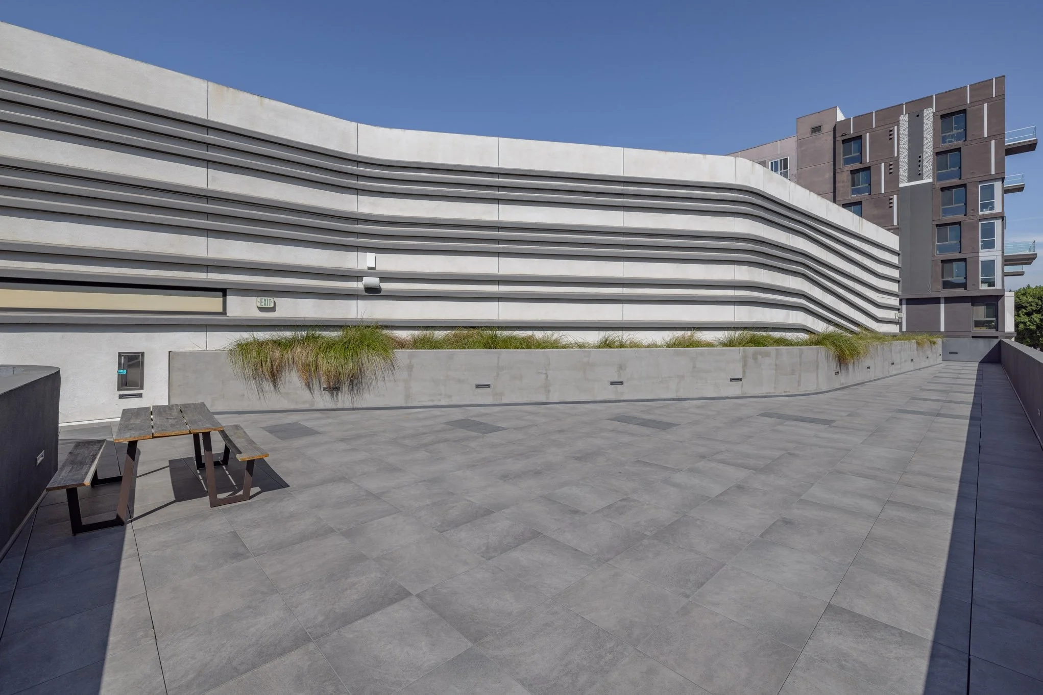 Modern urban rooftop terrace with gray tile flooring, a wooden picnic table, planter boxes with grasses, and a sleek building with horizontal lines against a clear blue sky.