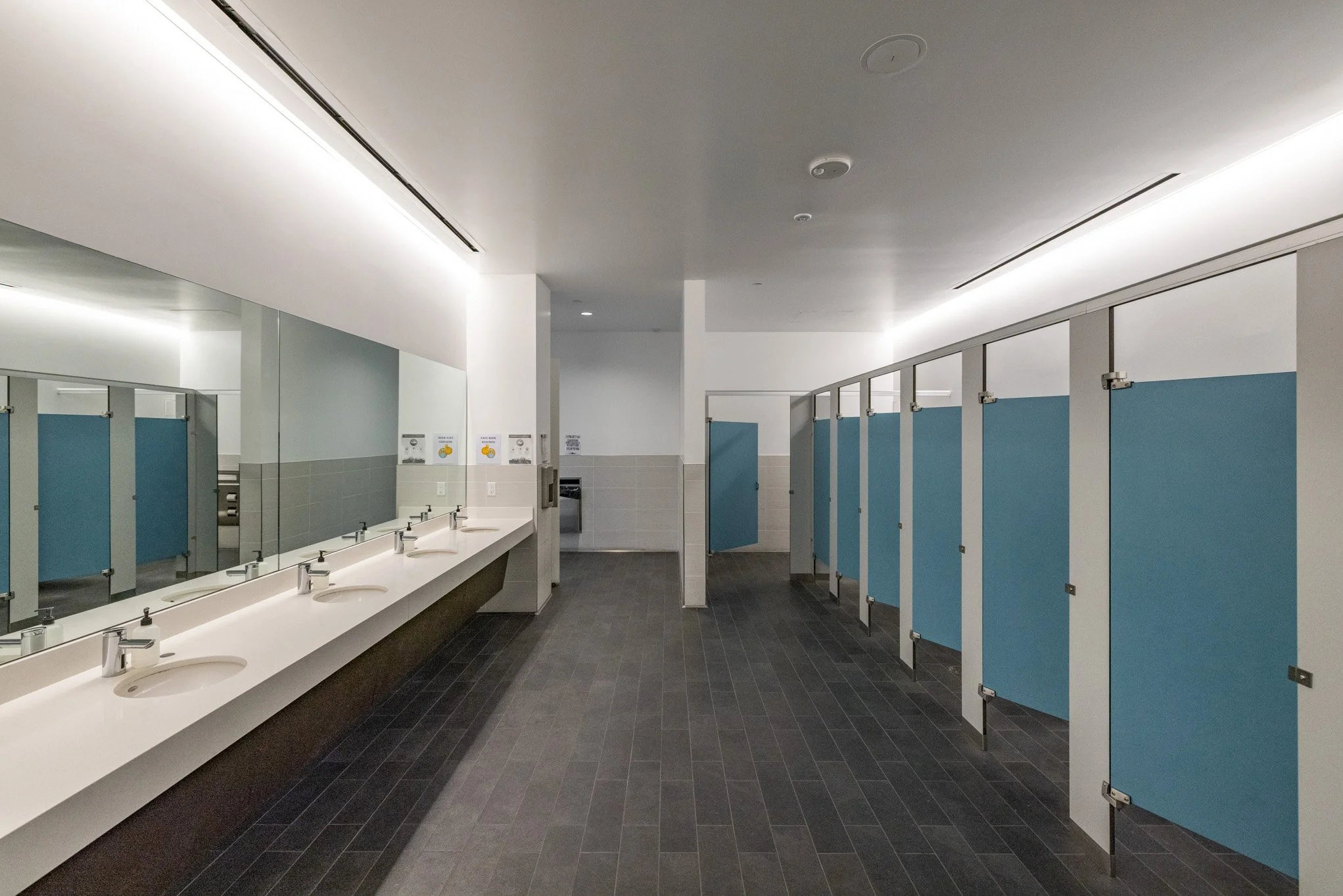 Empty school bathroom with four sinks, large mirror, hairdryer, and multiple unoccupied blue stall doors on dark tiled floor.