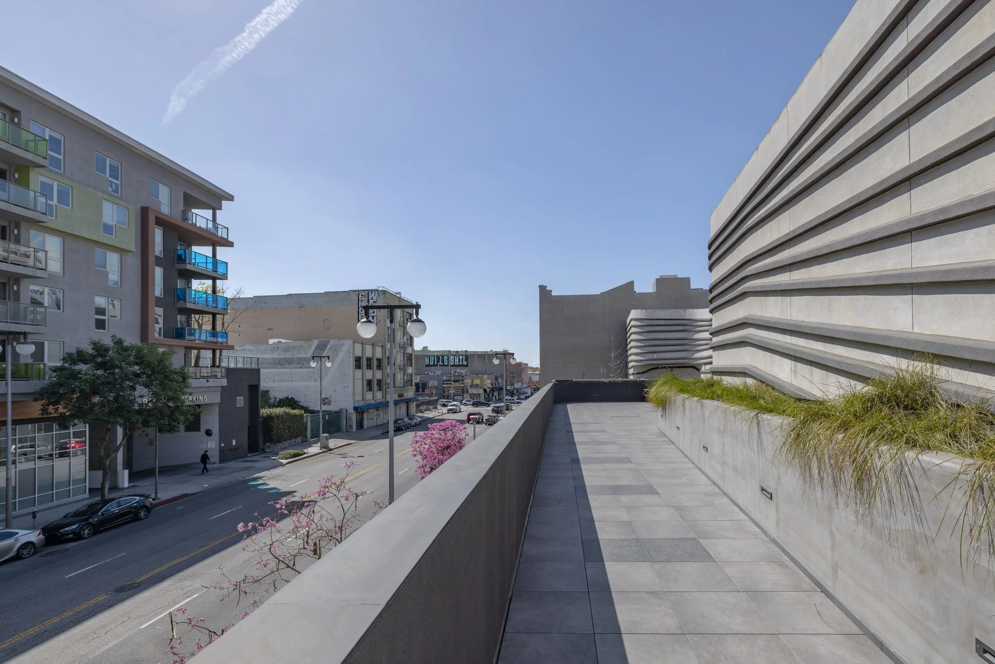 Empty balcony overlooking a city street with modern buildings, trees, parked cars, and a clear blue sky.