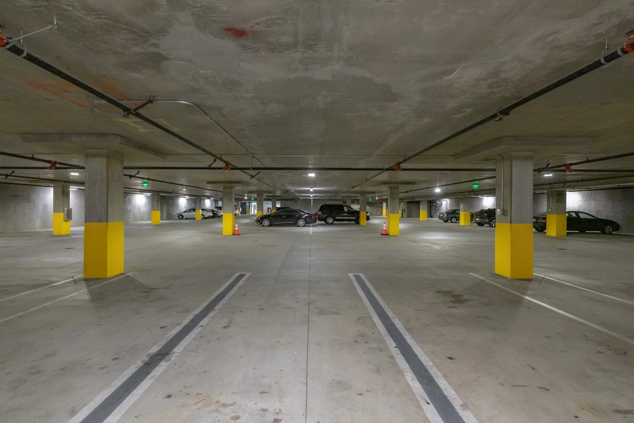 Empty underground parking garage with parked cars along the sides and yellow safety bumpers on concrete columns, illuminated by overhead lighting.