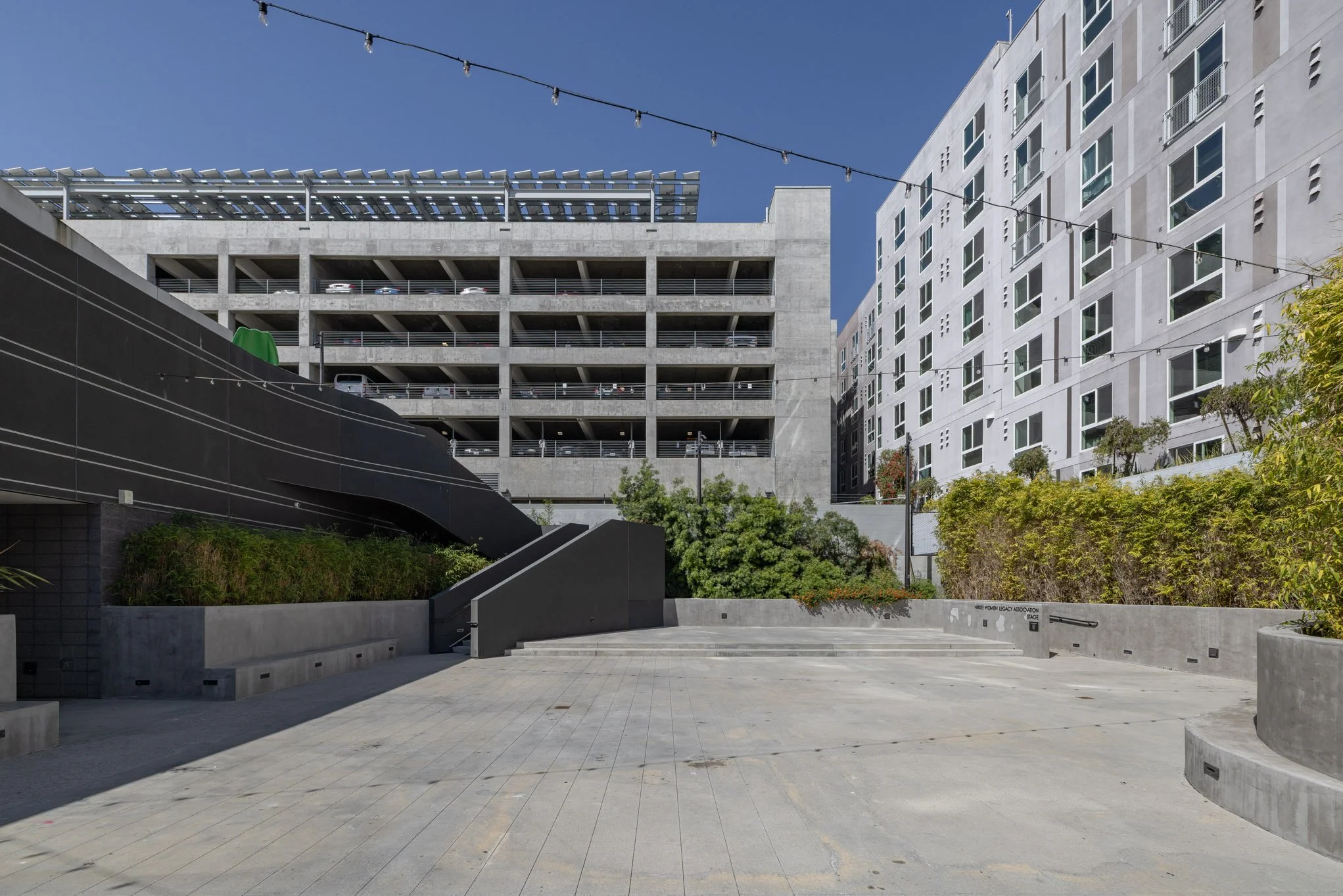 An urban outdoor courtyard area with concrete stairs and planters surrounded by modern multi-story residential buildings, with string lights overhead and a clear blue sky.