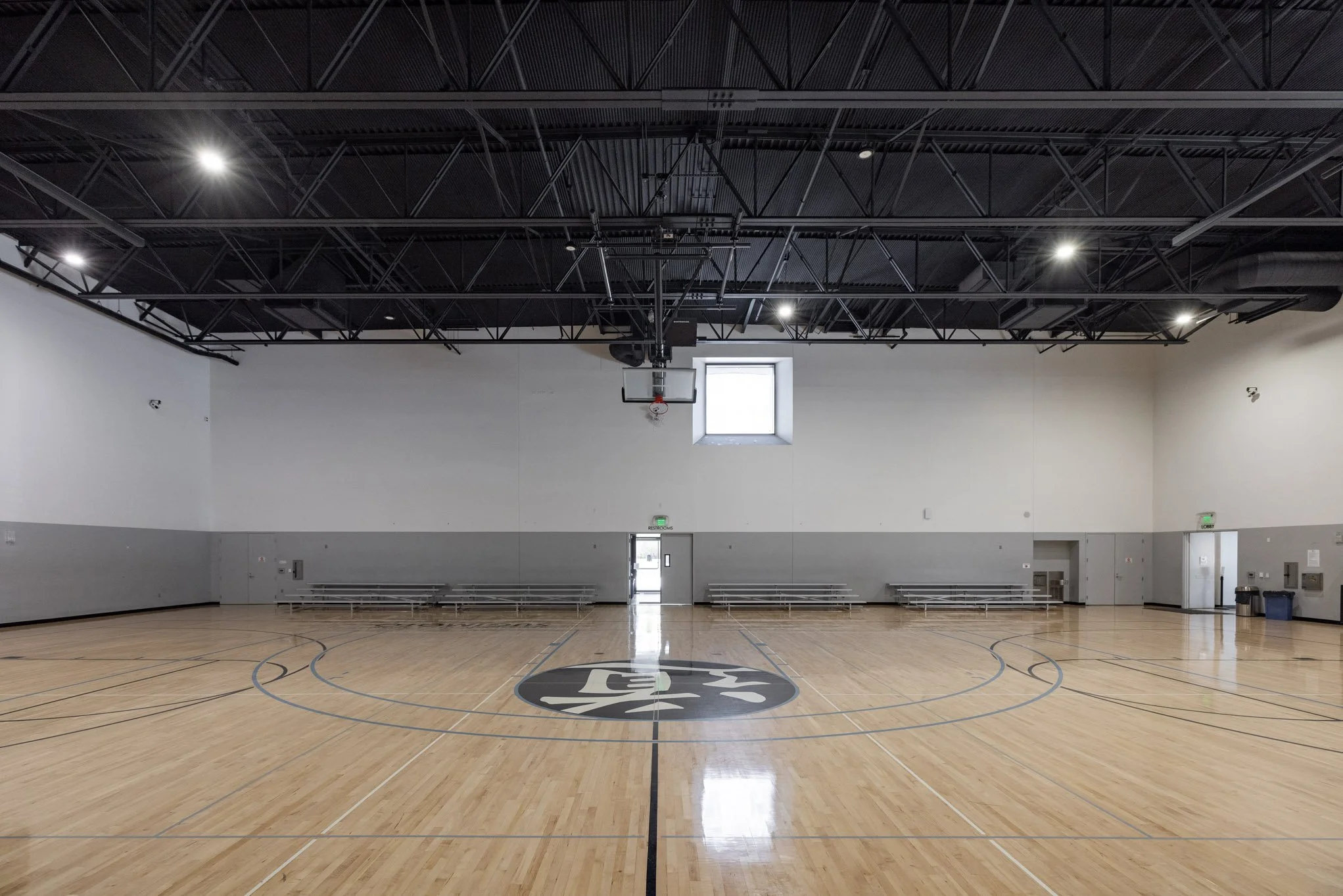 Empty indoor basketball court with a light wood floor, white walls, benches, a single window, and a basketball hoop.