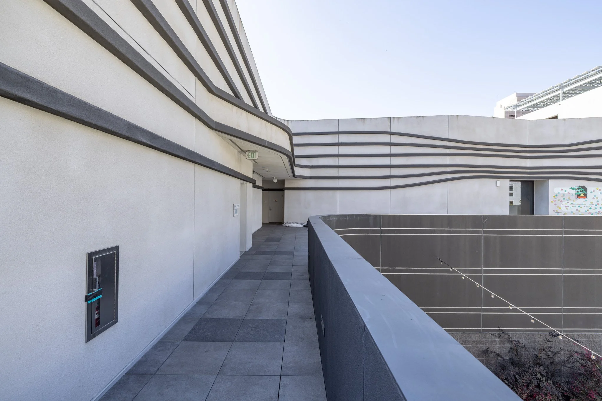 Empty rooftop walkway with white walls, black trim, exit sign, and a decorative mural in the distance.