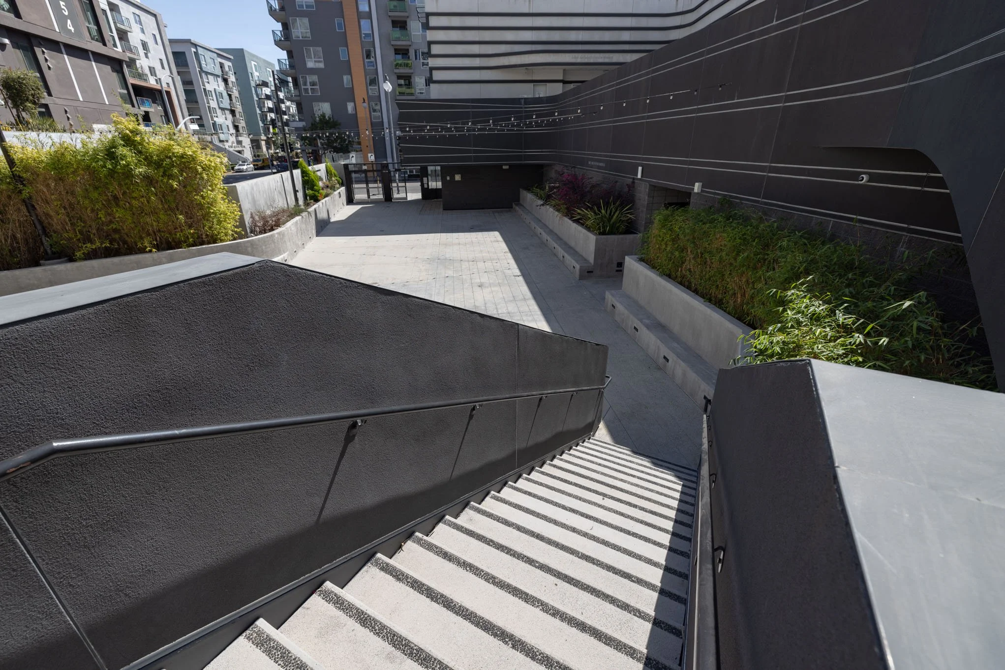 Outdoor stairs leading down to a modern urban courtyard with plants and high-rise buildings in the background.