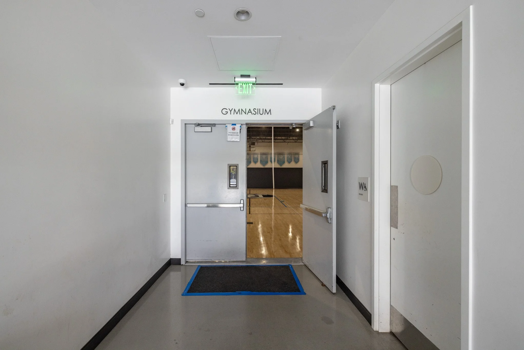 Entrance to a gymnasium with open double doors, white walls, black trim, and a wooden gymnasium floor visible inside.