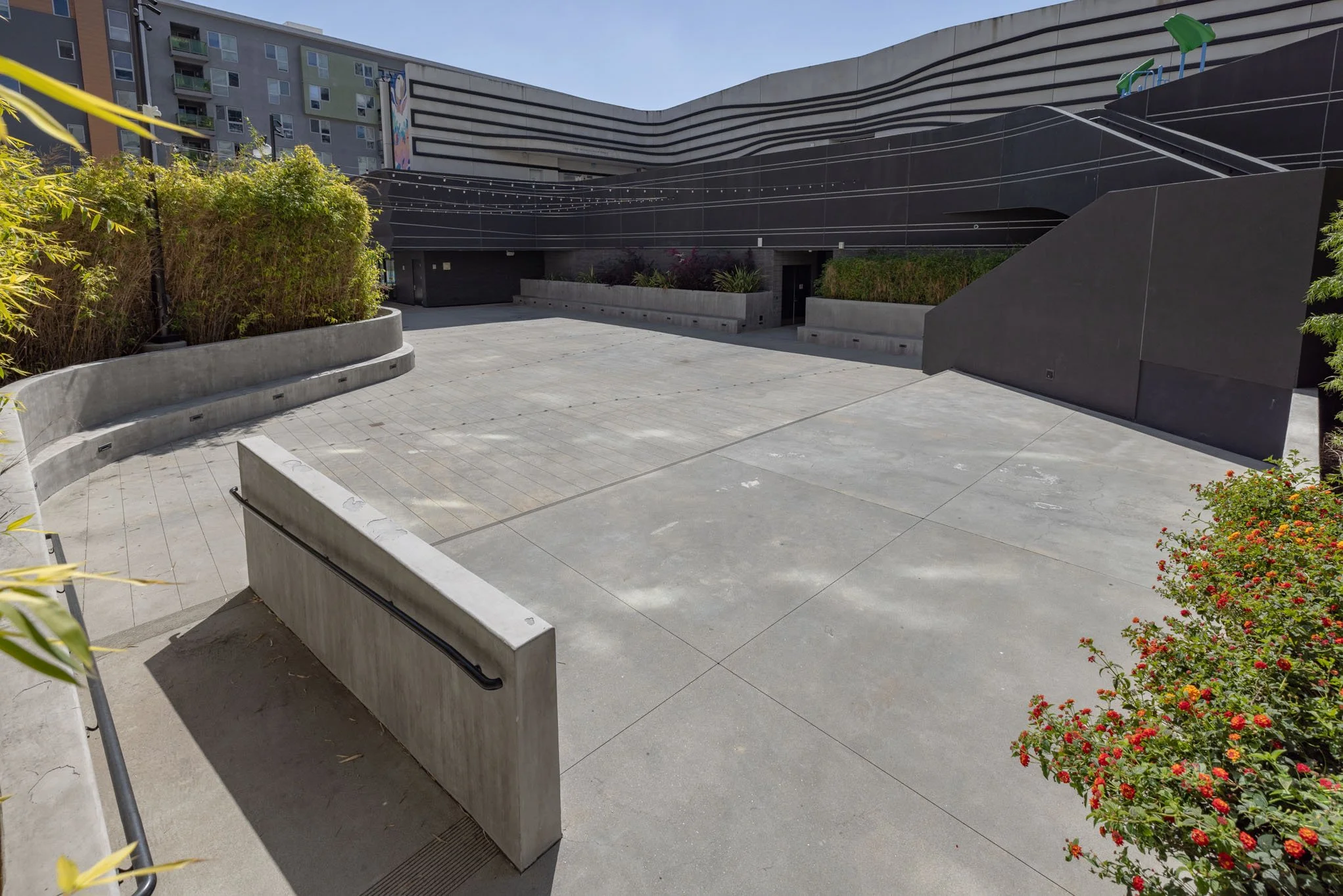 Empty outdoor urban space with concrete paving, planters with green bushes, and a modern black building with colorful murals in the background.