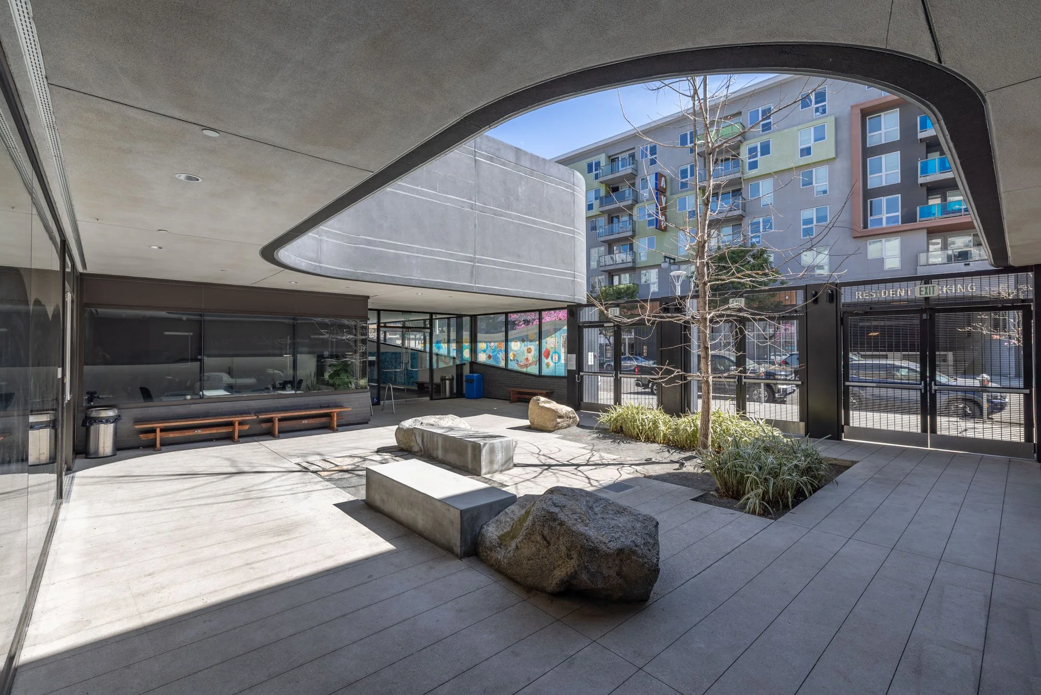 Modern apartment complex courtyard with concrete benches, small trees, and rocks, surrounded by glass and metal fences and a multicolored building in the background.