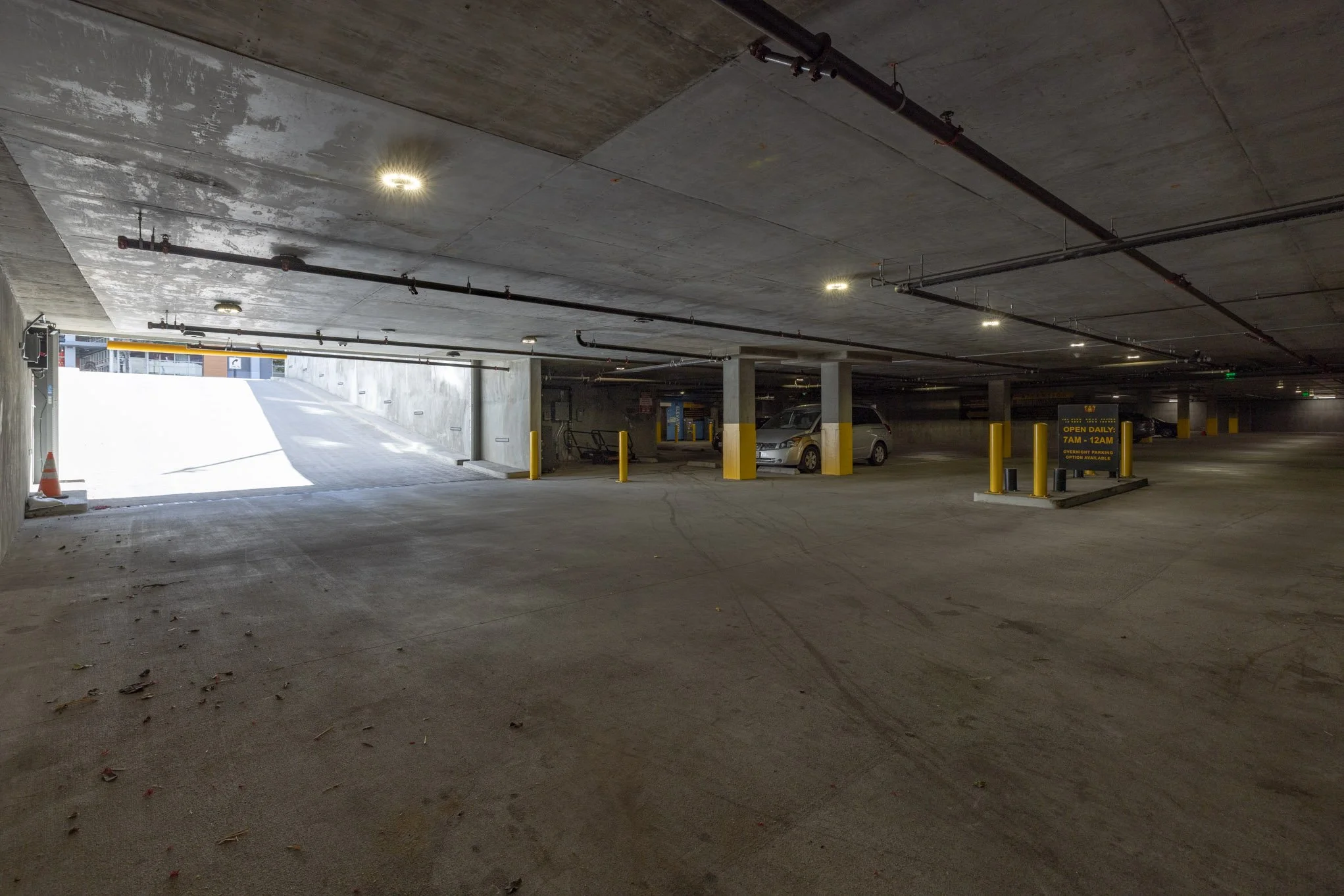 Underground parking garage with a few parked cars, concrete pillars painted yellow at the base, and a digital sign indicating parking hours, with an exit ramp in the background.