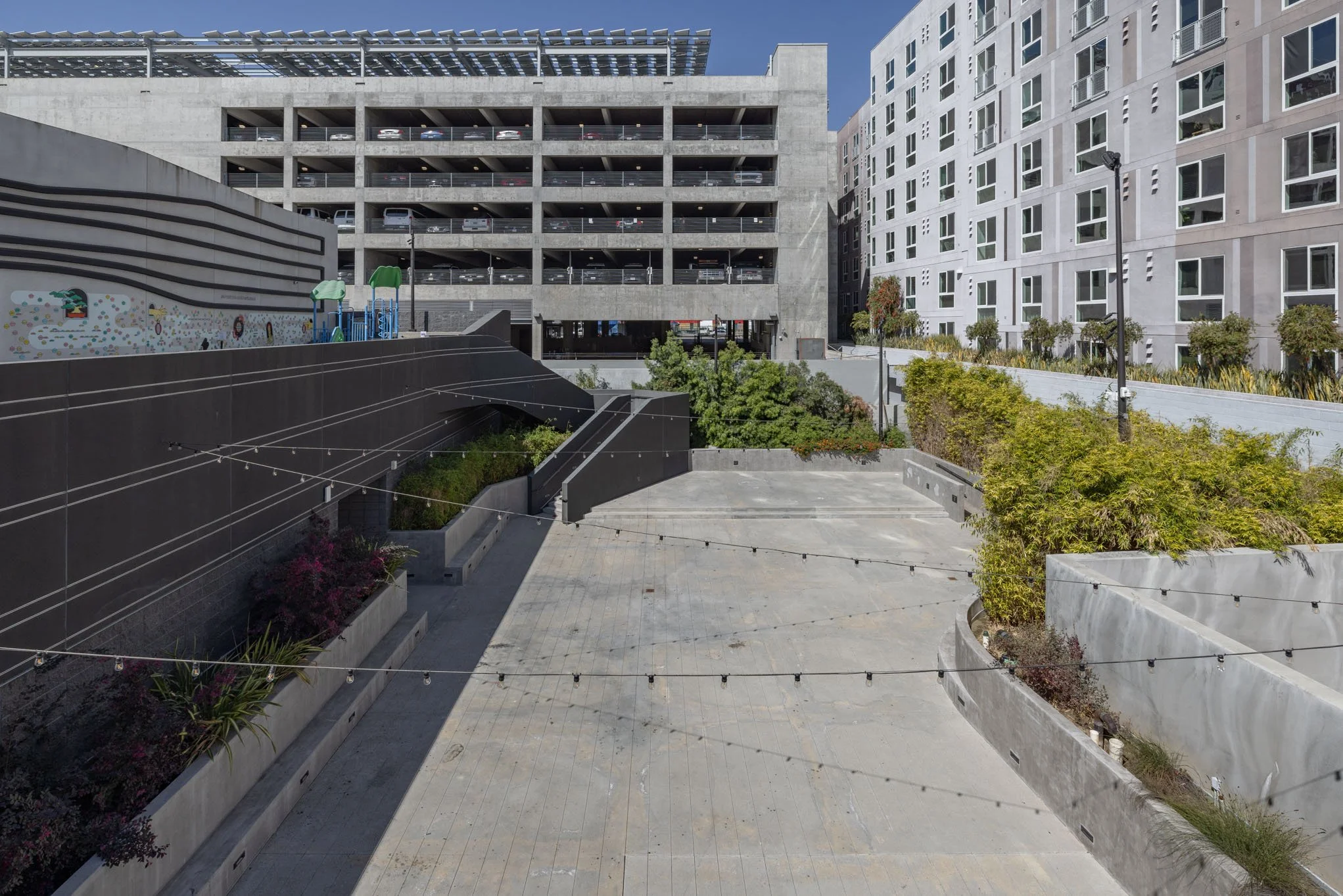 Modern cityscape featuring a multilevel parking garage, a small colorful playground, and an open concrete plaza surrounded by greenery and contemporary buildings.