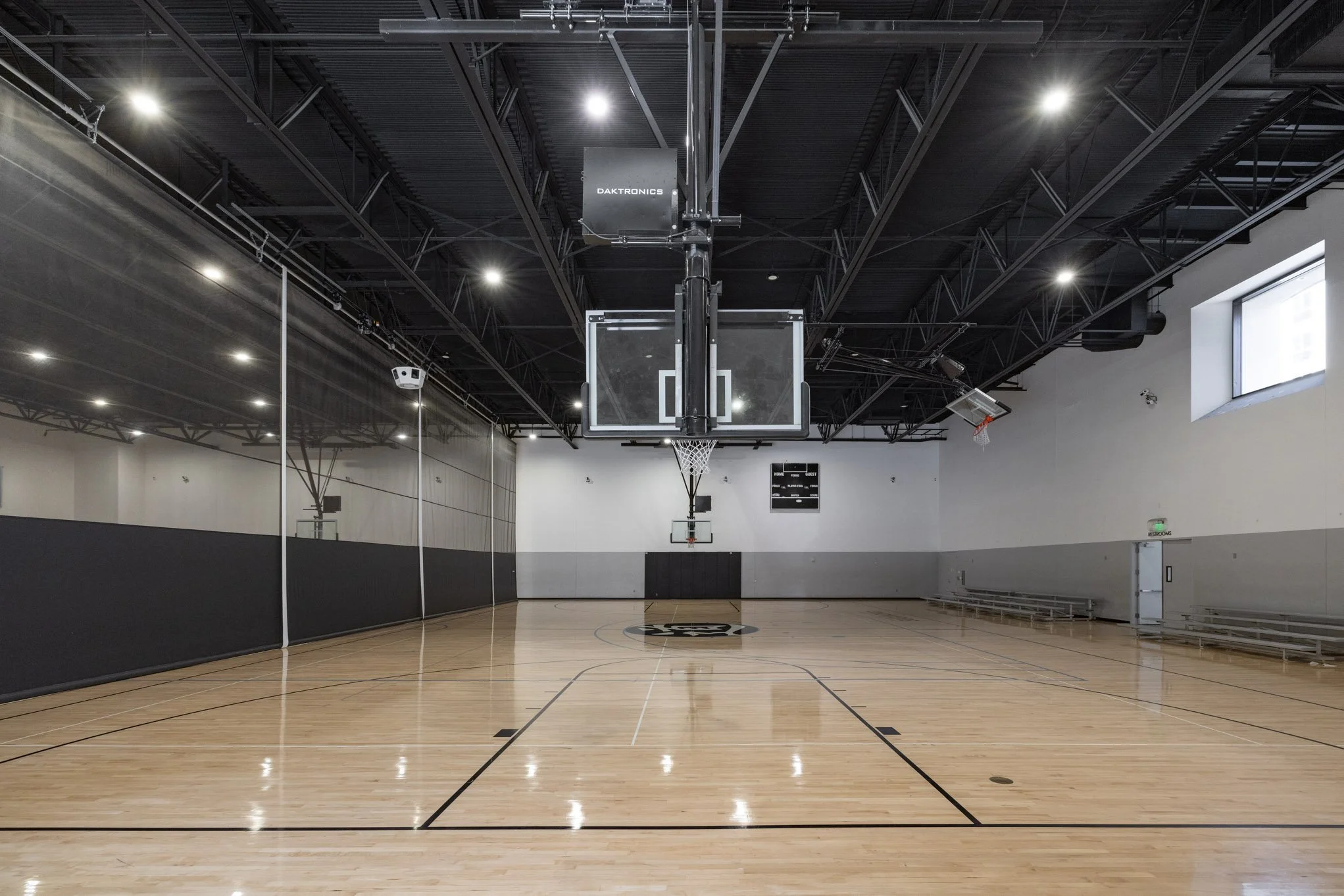 An empty indoor basketball gymnasium with a wooden floor, black and white walls, and two basketball hoops, one on each end.