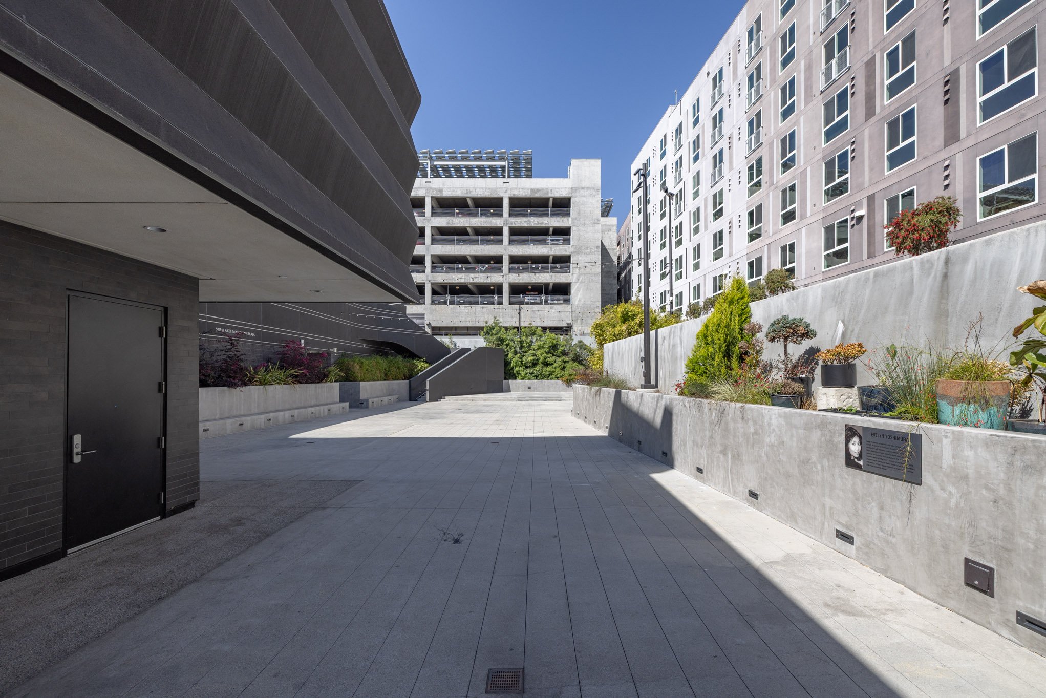 An urban outdoor courtyard area surrounded by modern multi-story buildings with potted plants on a concrete planter wall, under a clear blue sky.