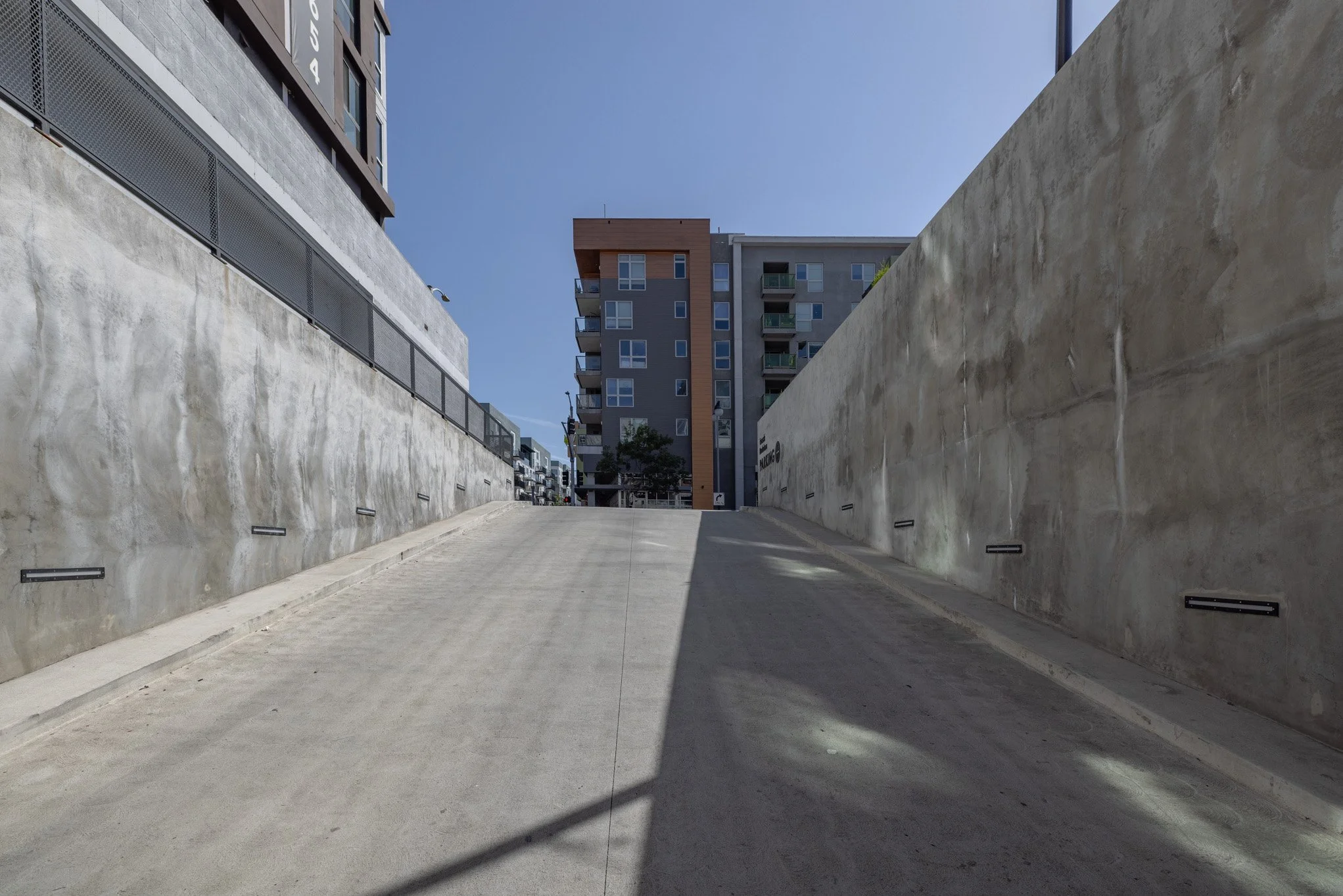 Uphill concrete driveway between high-rise buildings with modern apartments, a tree at the top, under a clear blue sky.