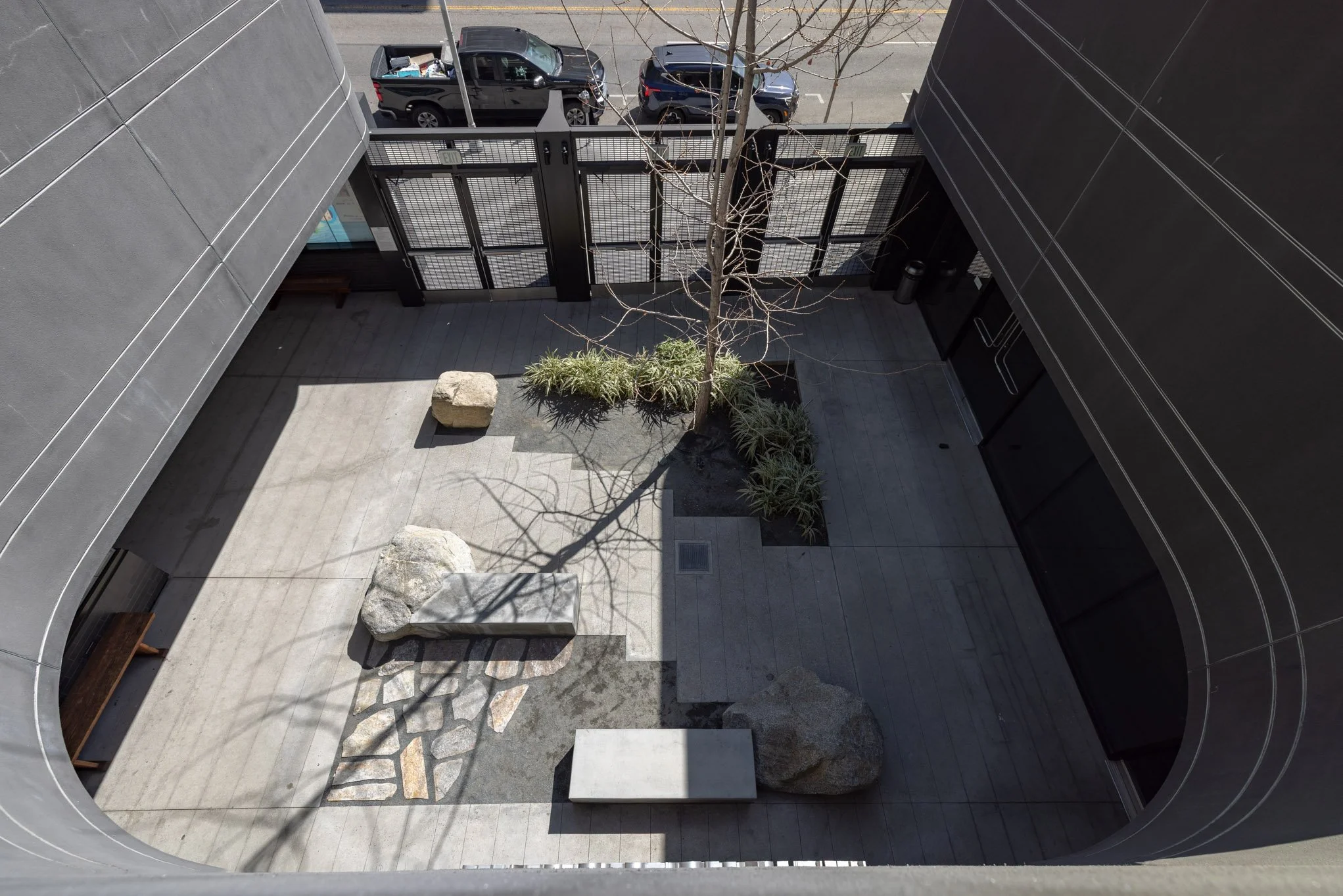 An outdoor courtyard with a leafless tree, a large rock, a bench, and a small landscaped area with plants, surrounded by a black metal fence and tall dark buildings, with parked cars visible beyond.