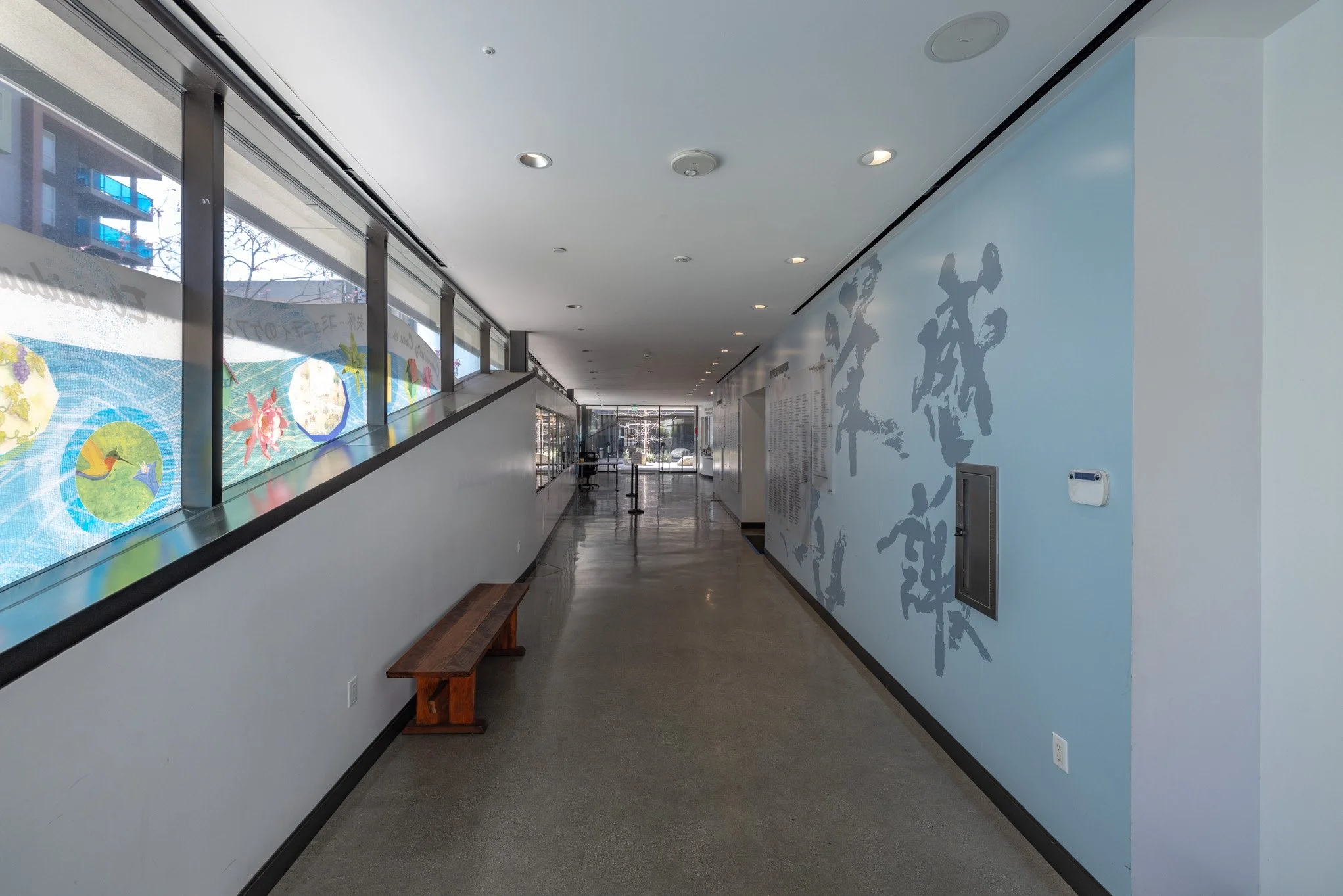 Empty hallway with large windows on the left side and a light blue wall with gray Asian characters on the right side.