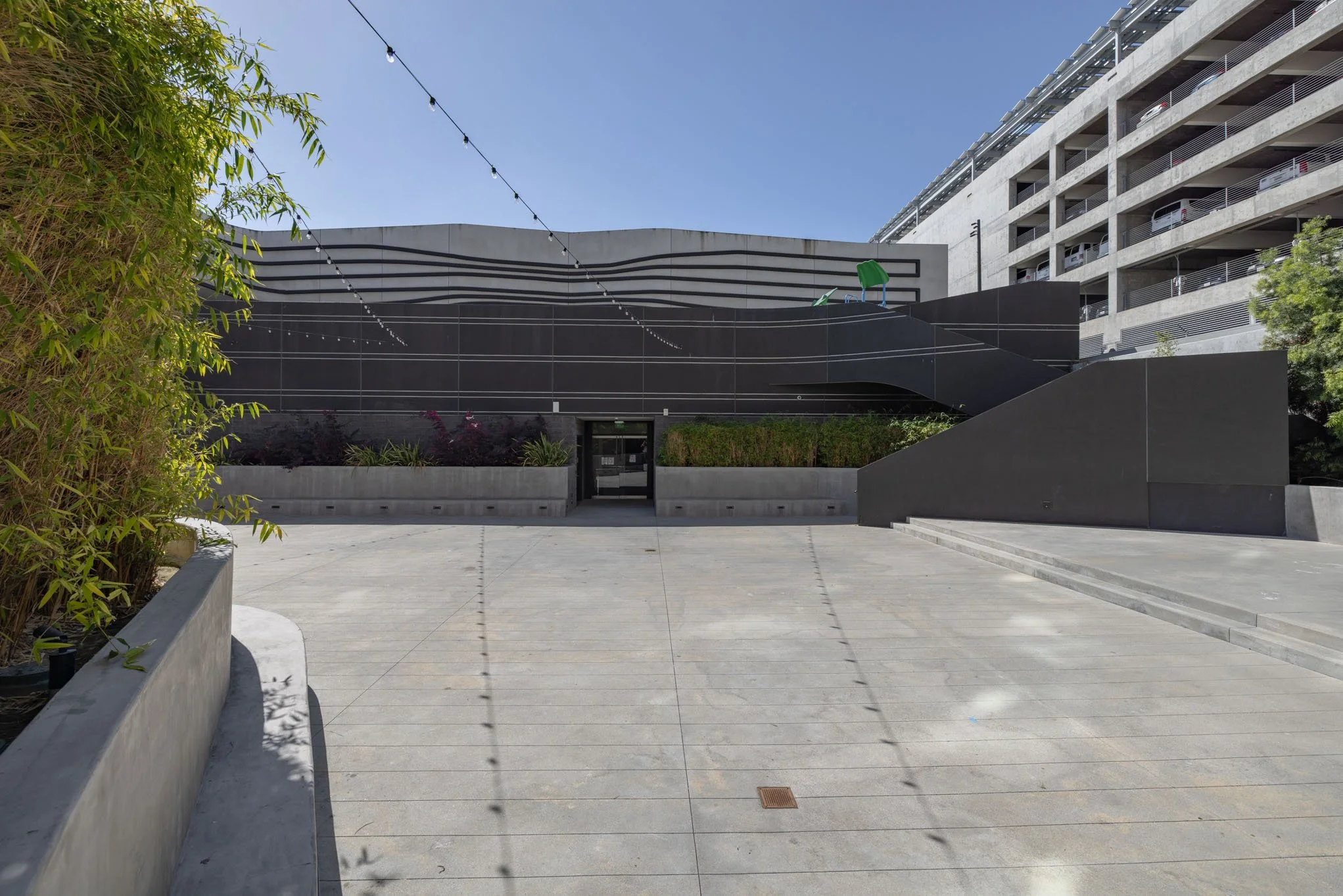 Modern black building with curved design, surrounded by concrete pavement, some planters with plants, and a high-rise building in the background, under a clear blue sky.