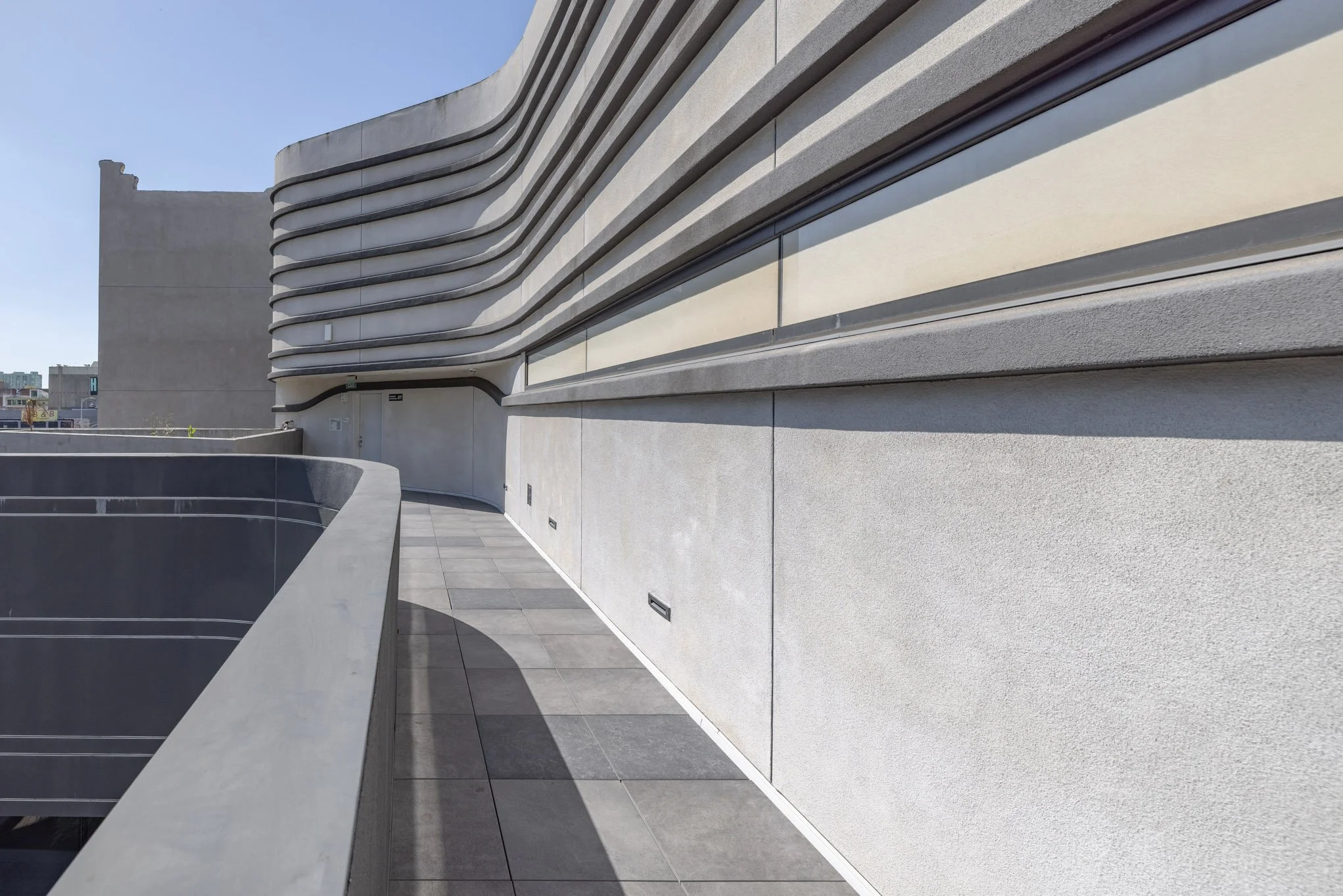 Modern building with curved architectural details, gray concrete walls, and a balcony with a gray railing, under a clear blue sky.