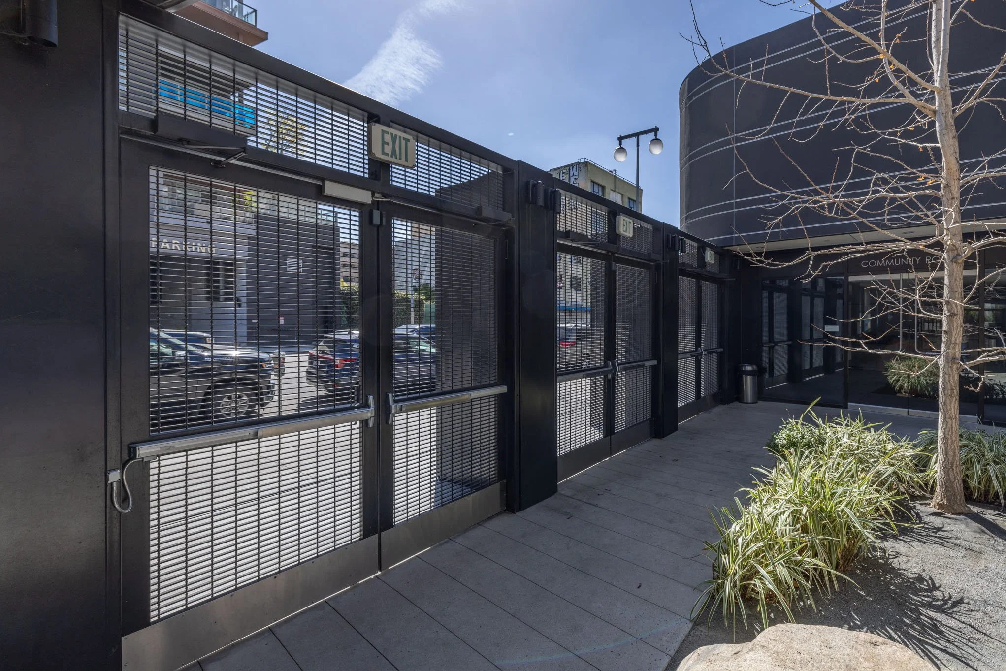 Black metal gate and entrance to a community area with trees and plants outside, parking lot visible through the gate, and a modern building in the background.
