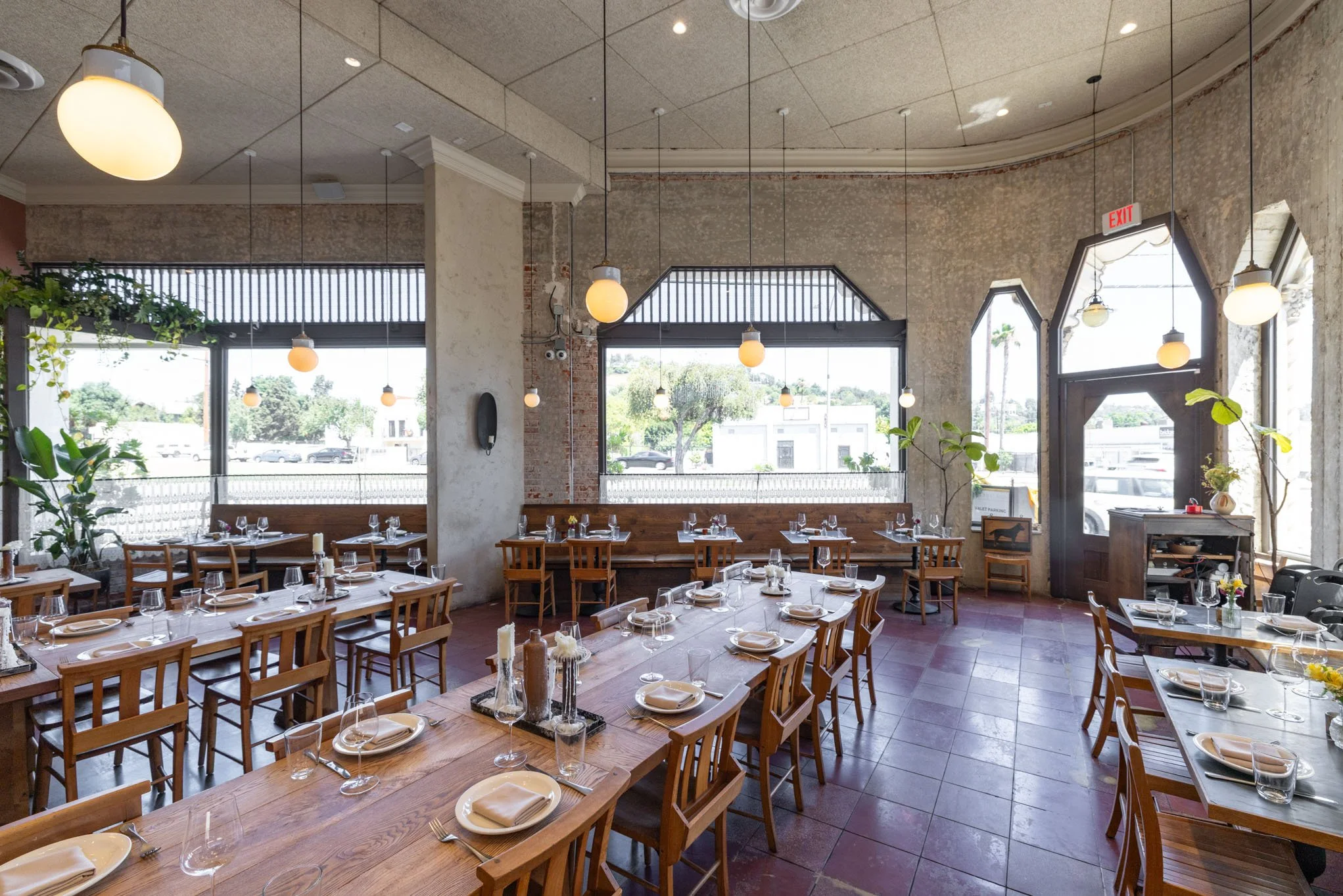 Interior of a restaurant with wooden tables and chairs, set with plates, glasses, and napkins, large windows with natural light, and green plants.