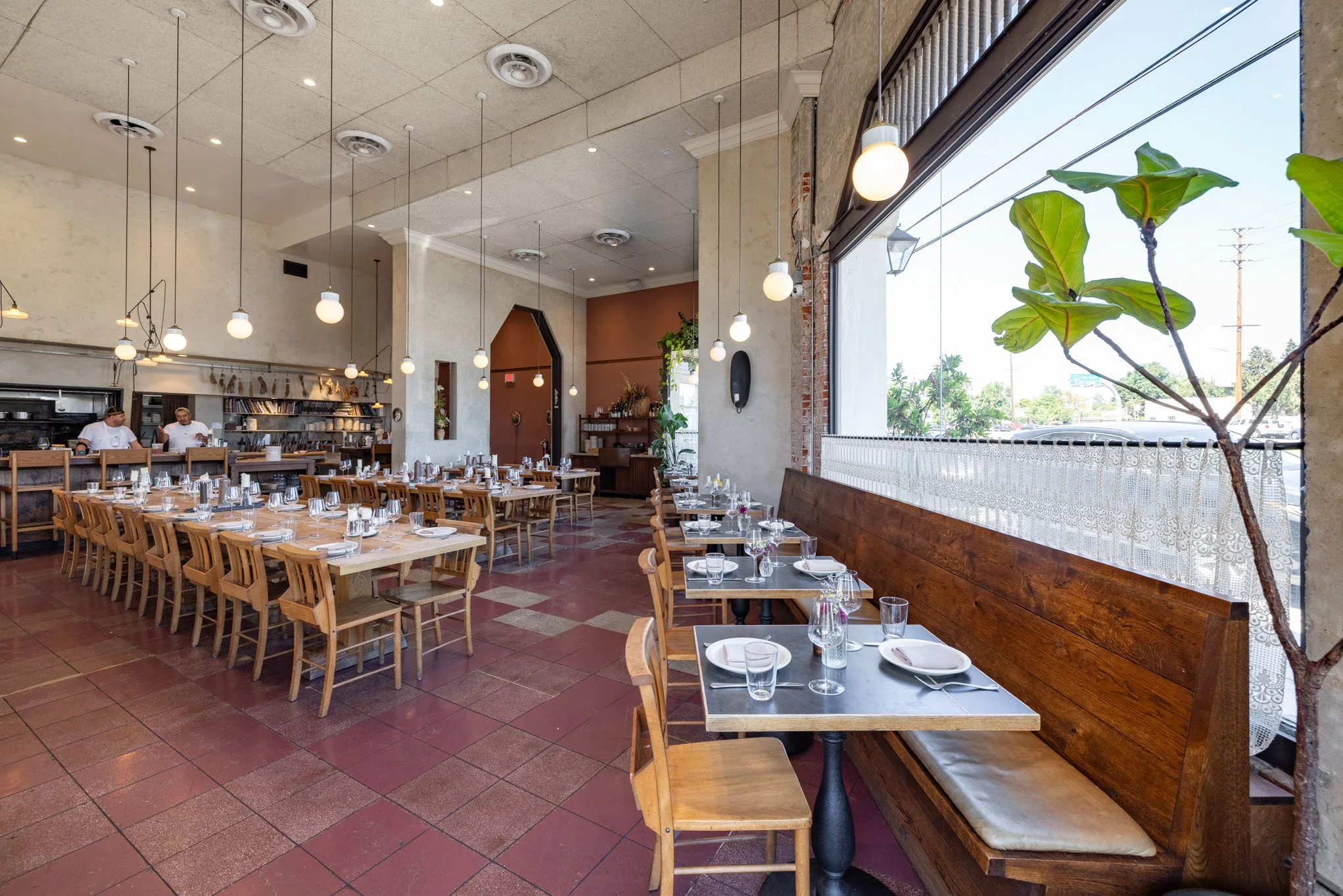 Interior of a restaurant with wooden chairs and tables set with glassware and plates, large window with lace curtains, hanging lights, and chefs in the kitchen area.