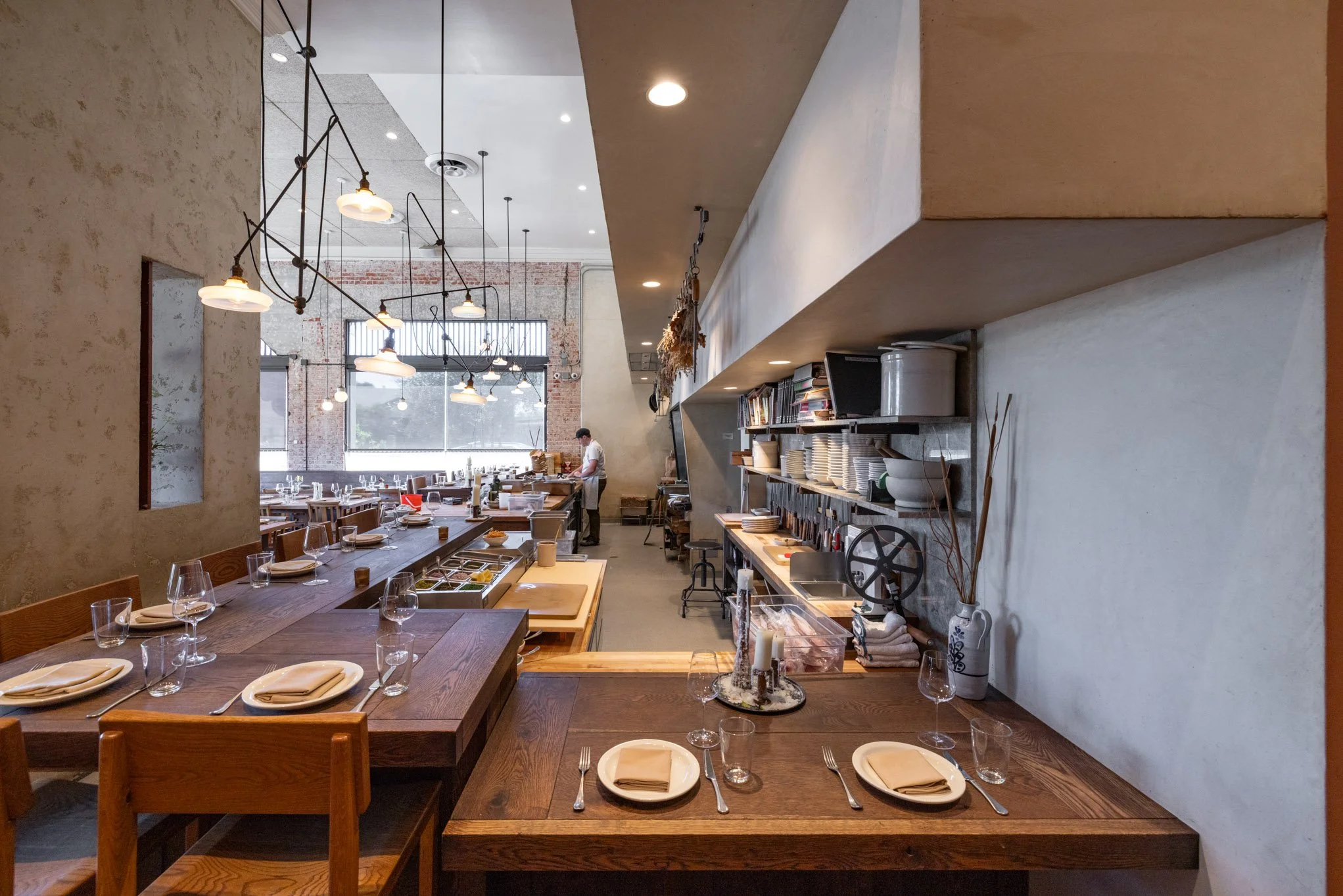 Interior of a modern restaurant with wooden tables set with plates, glasses, and napkins, open kitchen area with shelves and kitchen tools, and a large window letting in natural light.