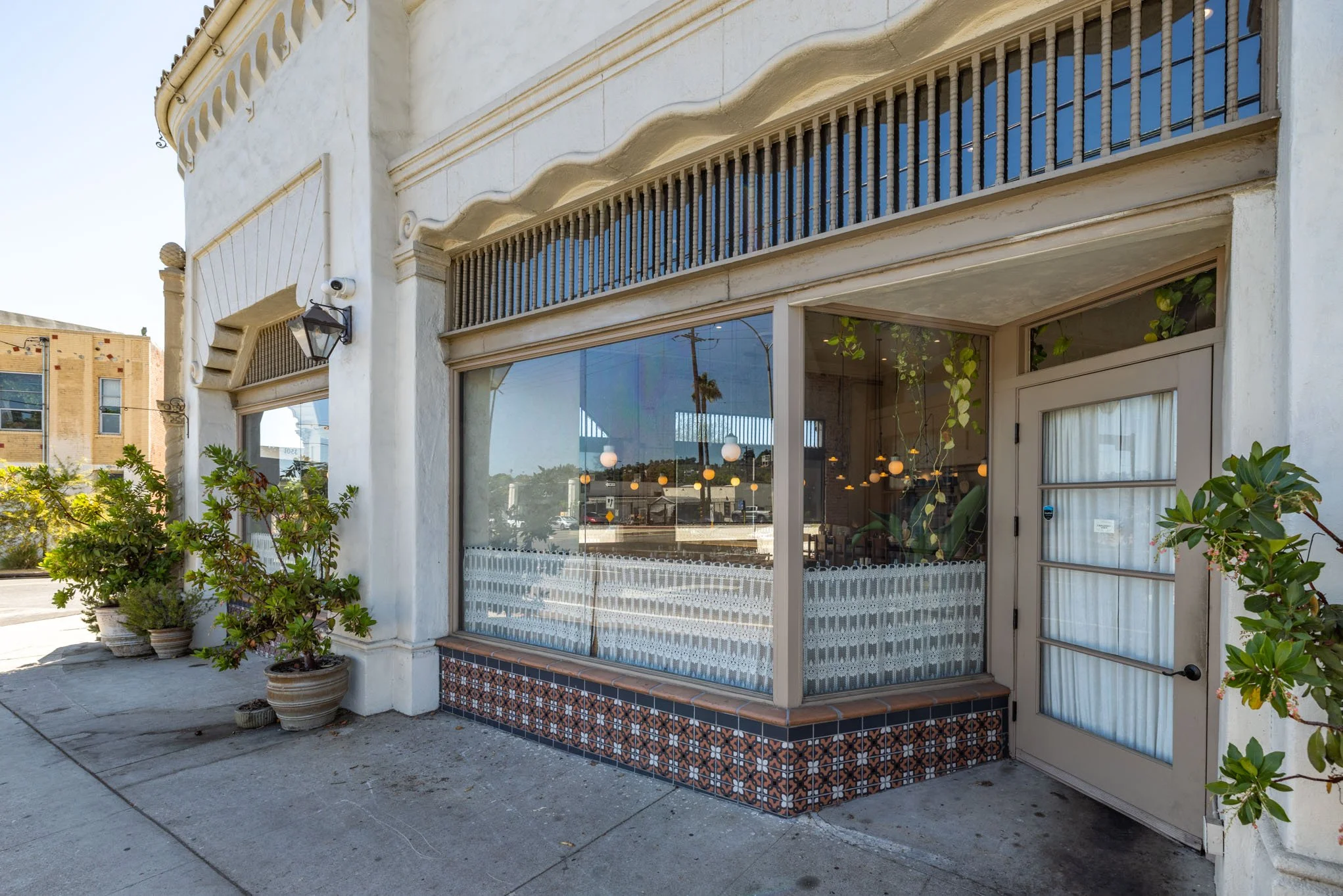 Exterior view of a storefront with large glass windows, potted plants outside, and a decorative tiled base around the corner, with reflection of street and streetlights in the window.