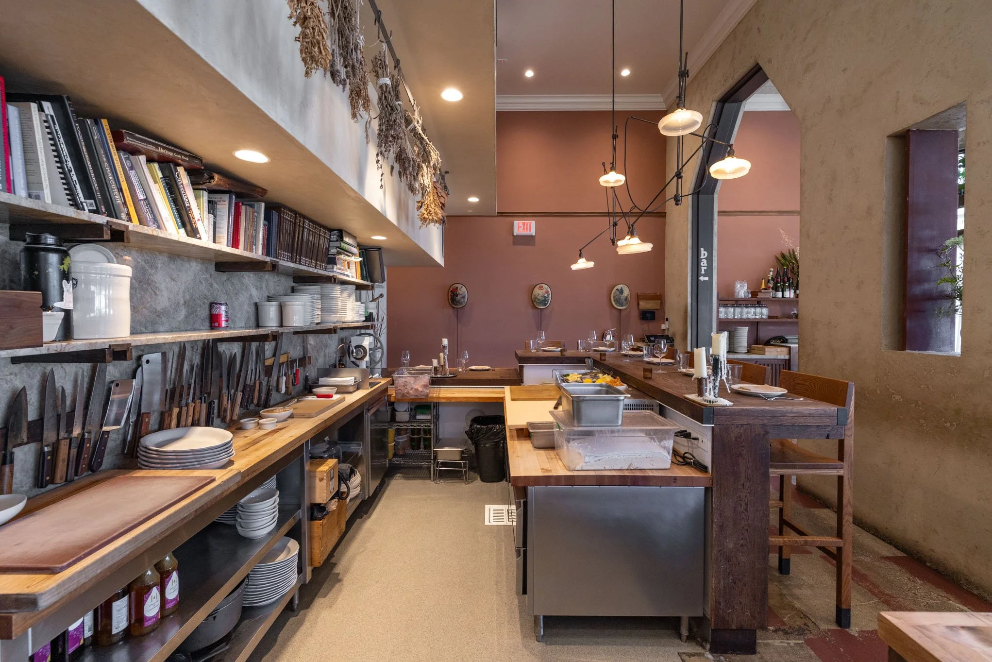 Interior of a restaurant kitchen with countertops, shelves filled with cookbooks, kitchen utensils, and plates, and tables set with wine glasses and candles.