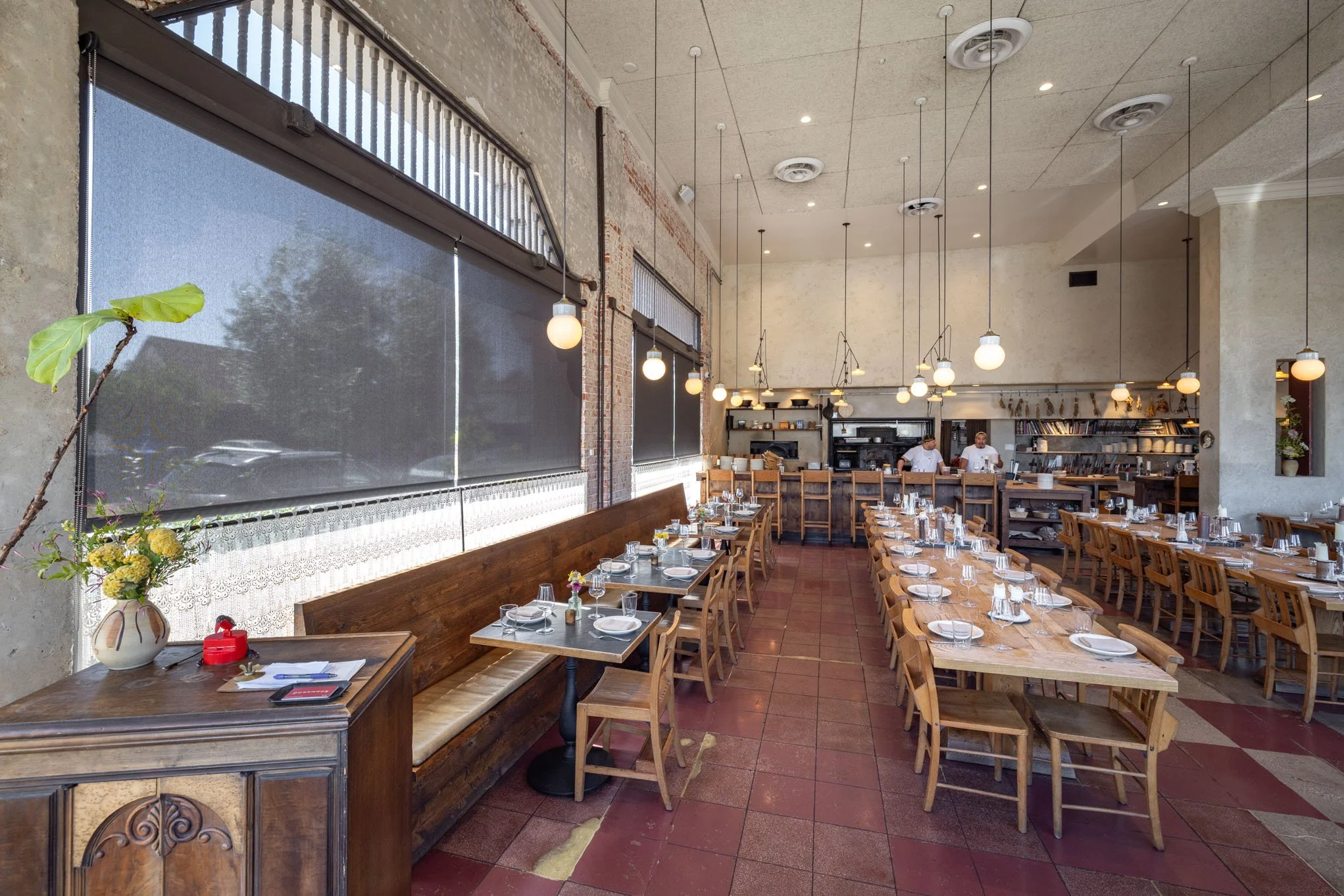 Empty restaurant dining area with wooden chairs and tables set for service, large windows with blinds, and hanging pendant lights, with two staff members in the background.