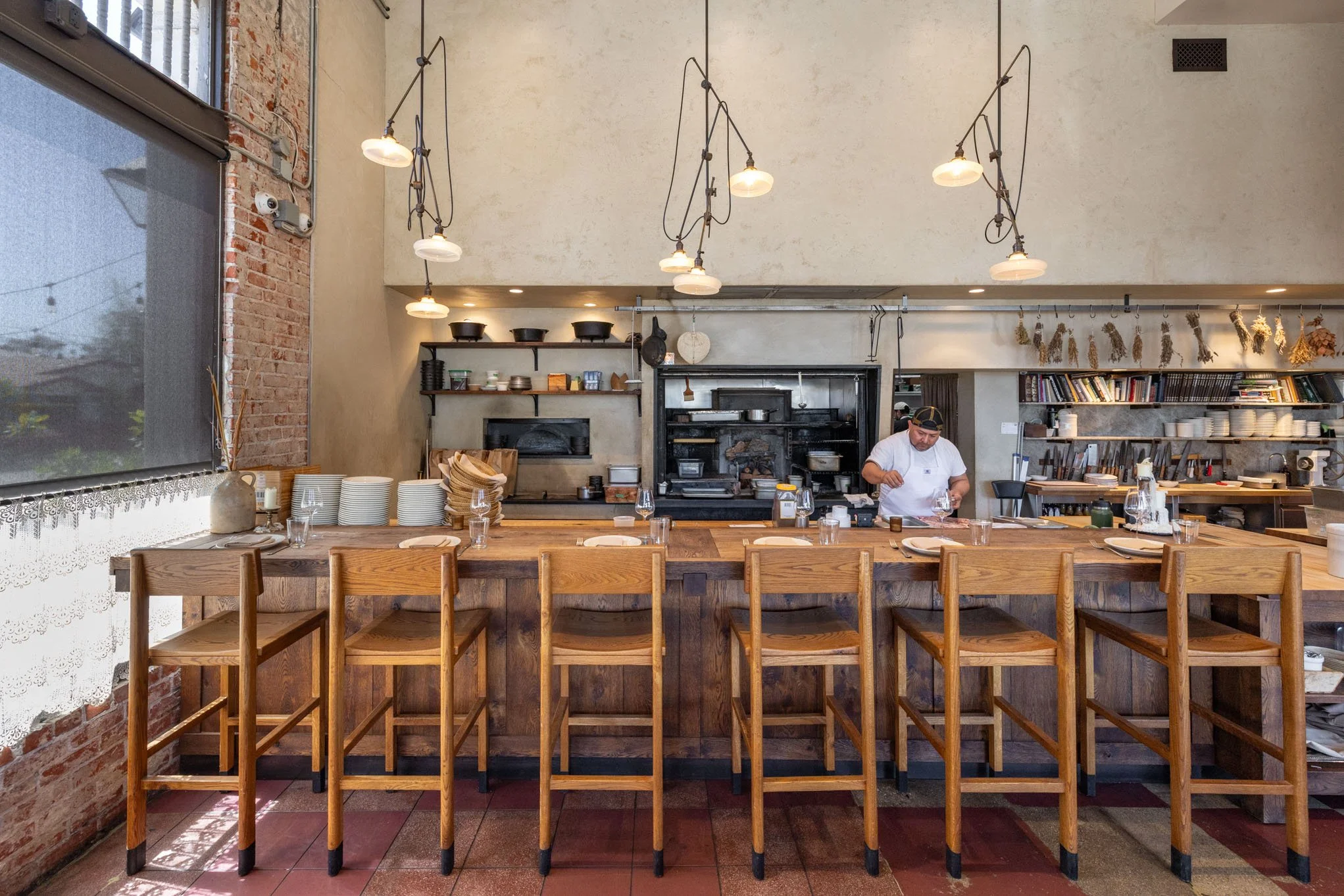 Inside a cozy restaurant with a long wooden counter, a chef preparing food, hanging pendant lights, and shelves with kitchenware and books.