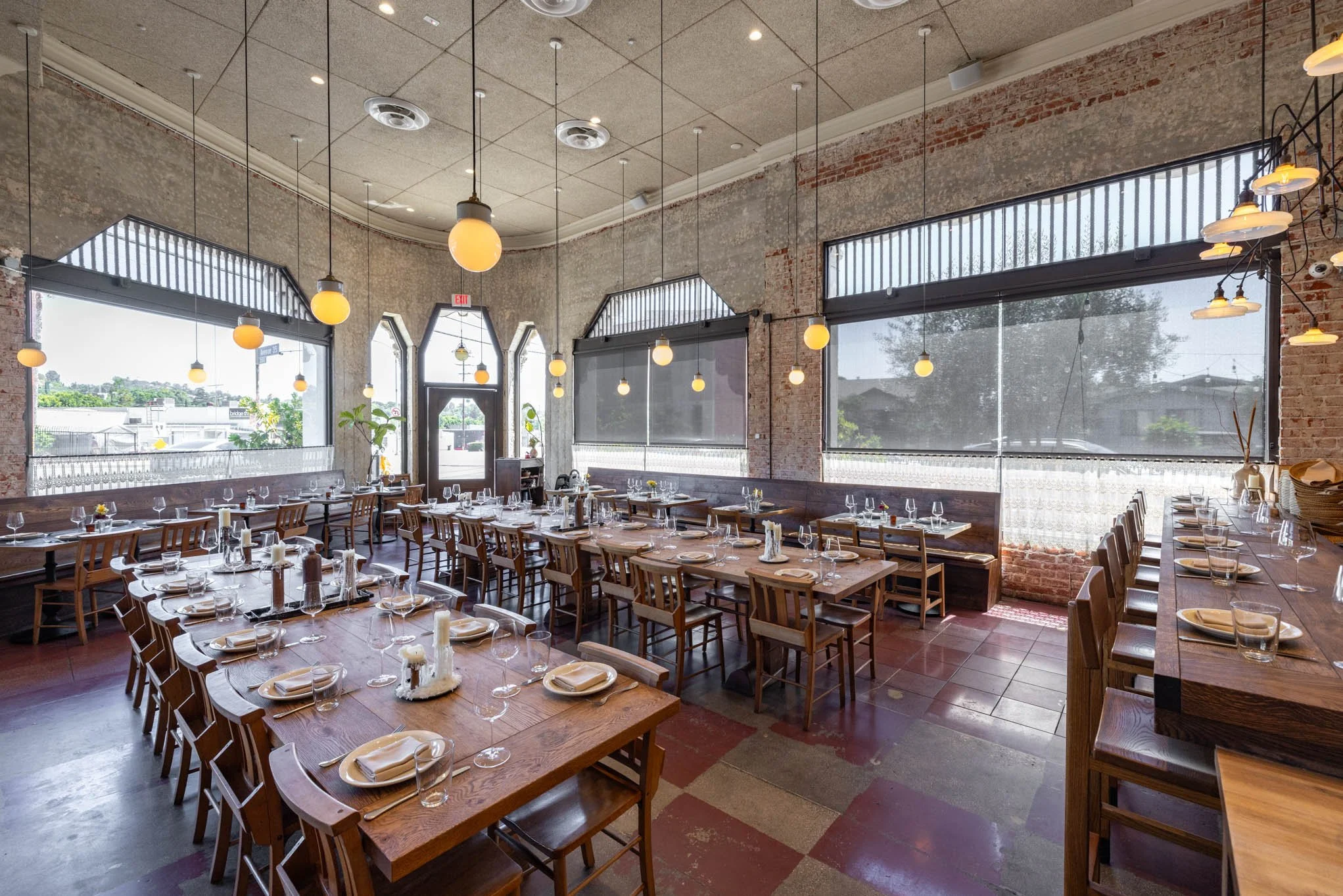 Interior of an empty restaurant with rustic brick walls, large windows with blinds, multiple dining tables set with plates, glasses, napkins, and candles, and warm hanging pendant lights.
