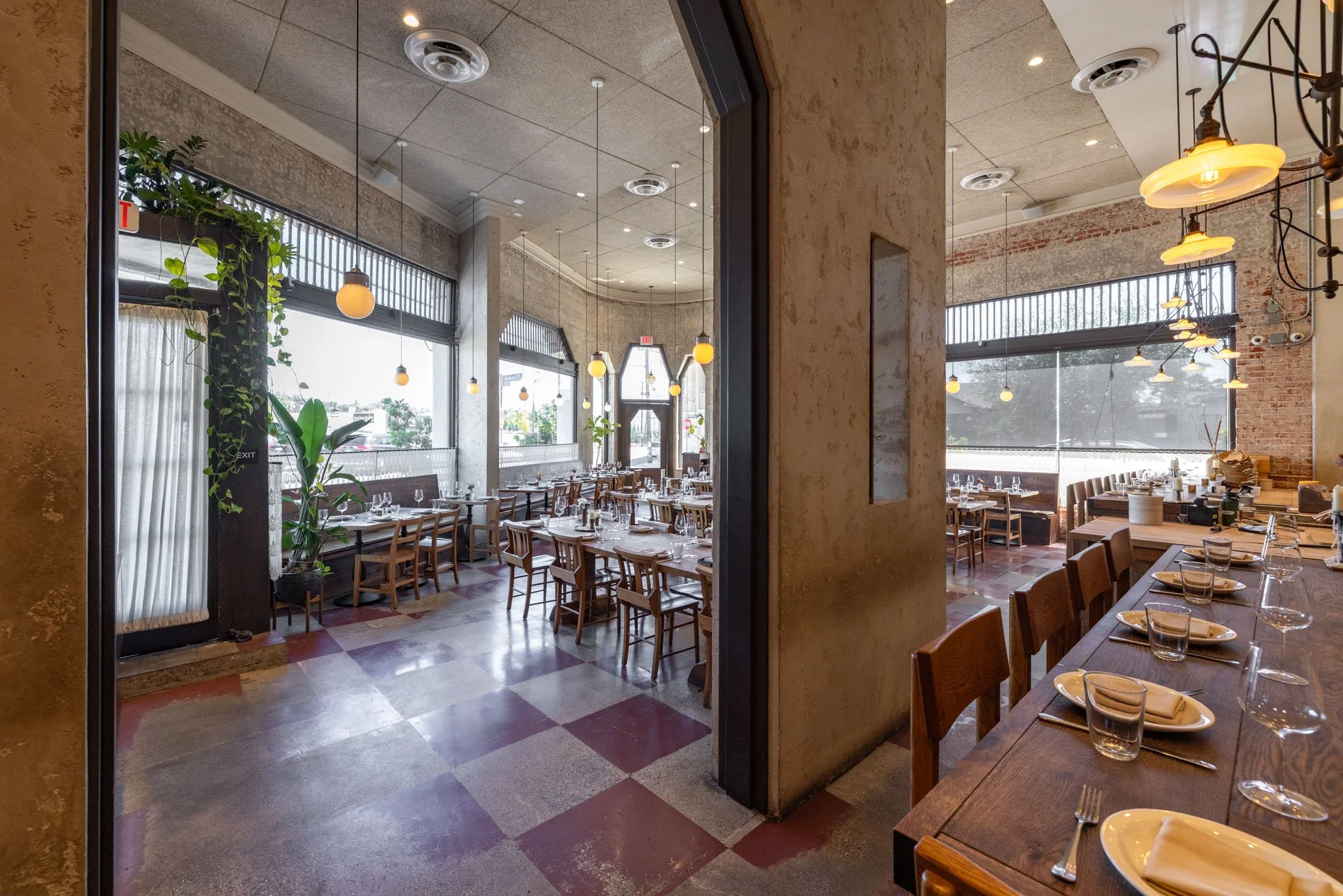 Interior view of a restaurant dining area with wooden tables and chairs, large windows, and warm pendant lighting.