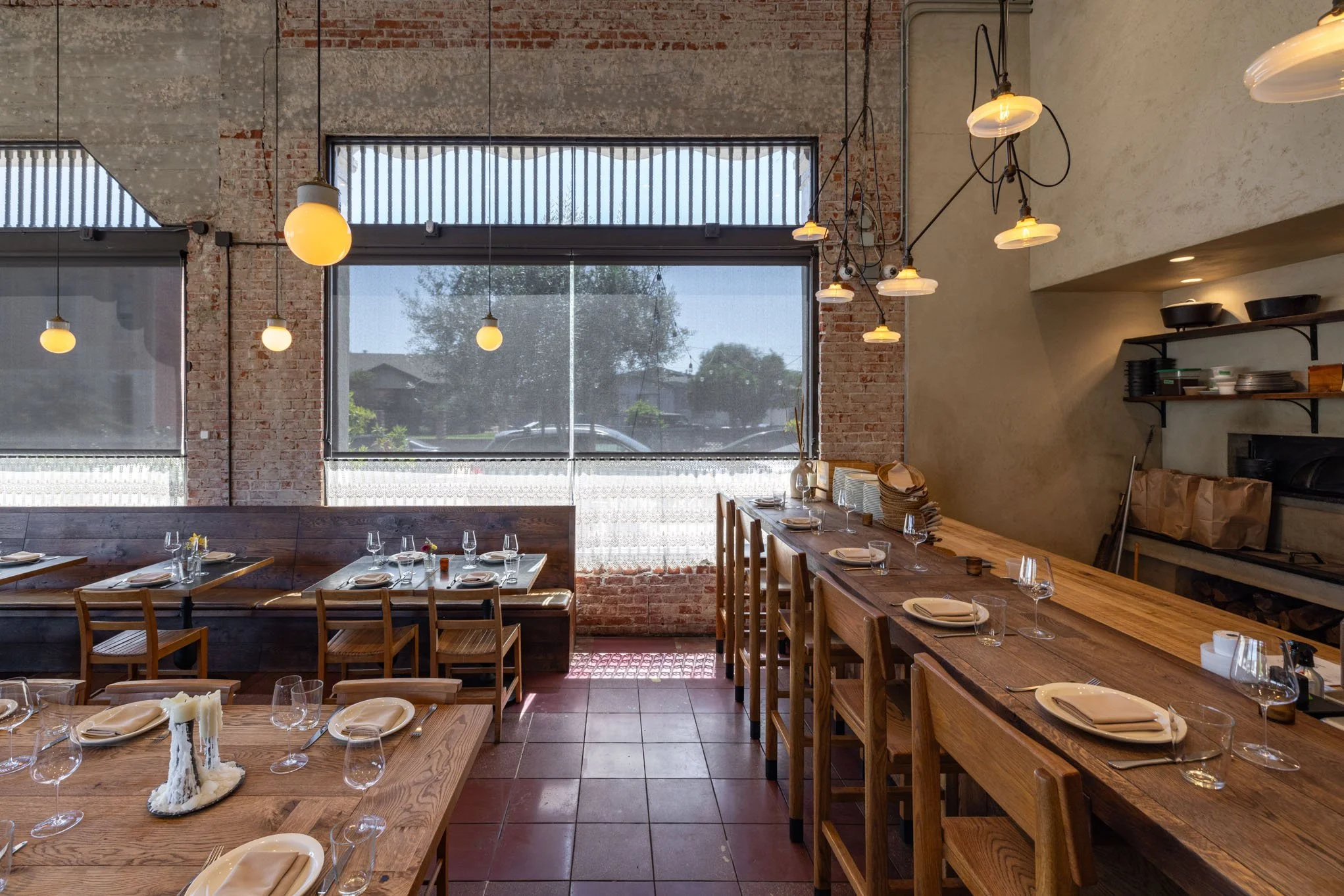 Interior of a modern restaurant with wooden tables, chairs, and hanging lights, large windows revealing a sunny outdoor scene.