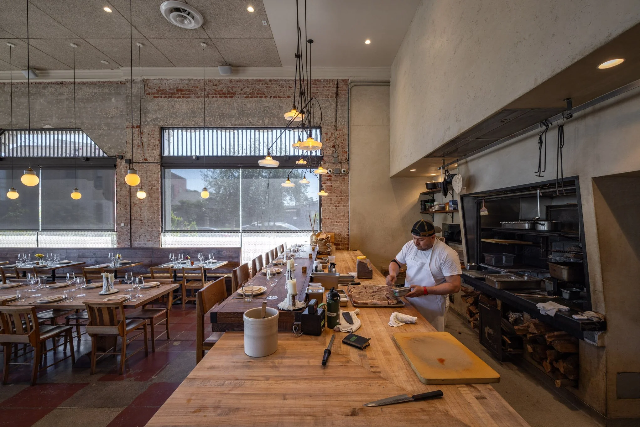 A chef preparing food in an open kitchen at a restaurant with tables set for guests, large windows, and modern hanging lights.