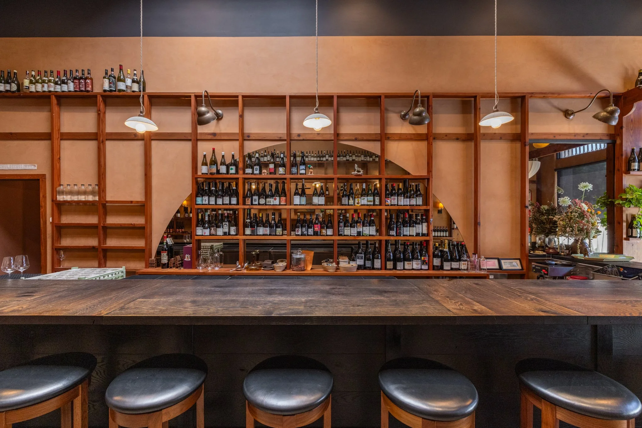 Empty bar counter with a wooden back shelf filled with wine bottles, surrounded by bar stools in a cozy restaurant setting.