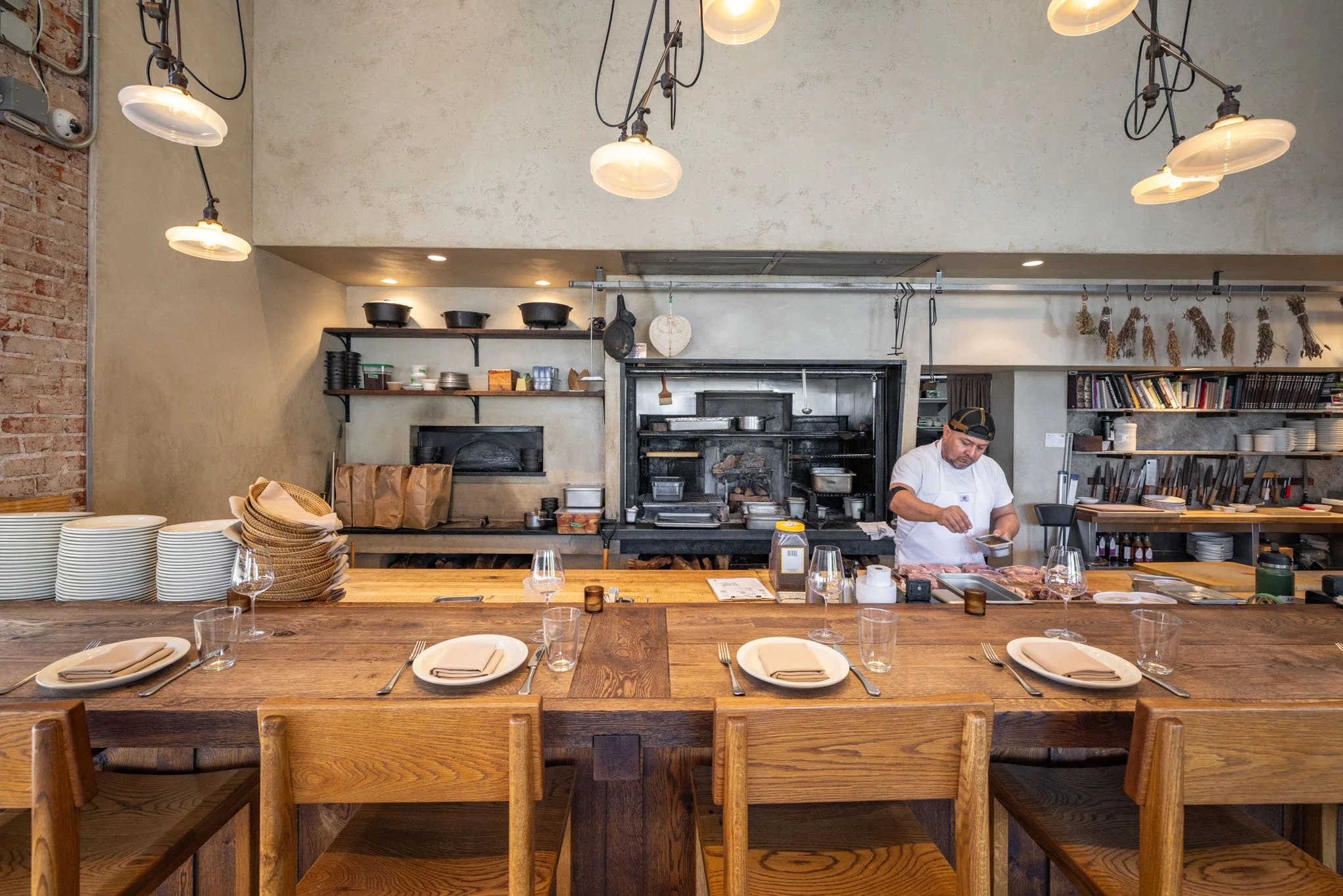 Interior of a restaurant kitchen with a chef preparing food, set tables with plates, napkins, glasses, and silverware, wooden chairs and countertops, shelves with various kitchen items, and industrial lighting.