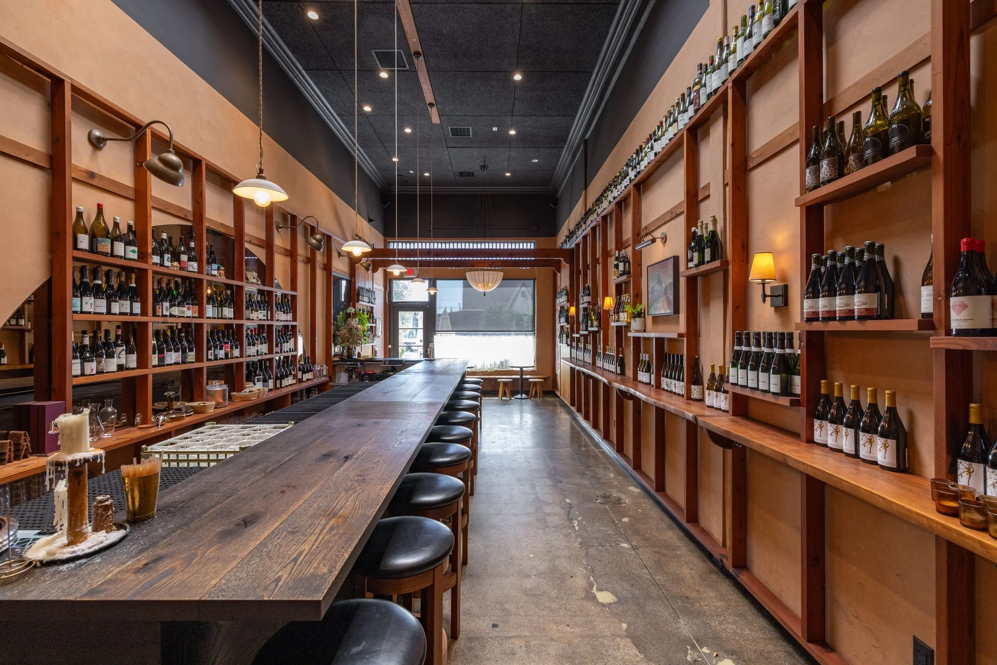 Interior of a wine bar or shop with a long wooden counter and shelves filled with wine bottles, warm lighting, and minimal decor.