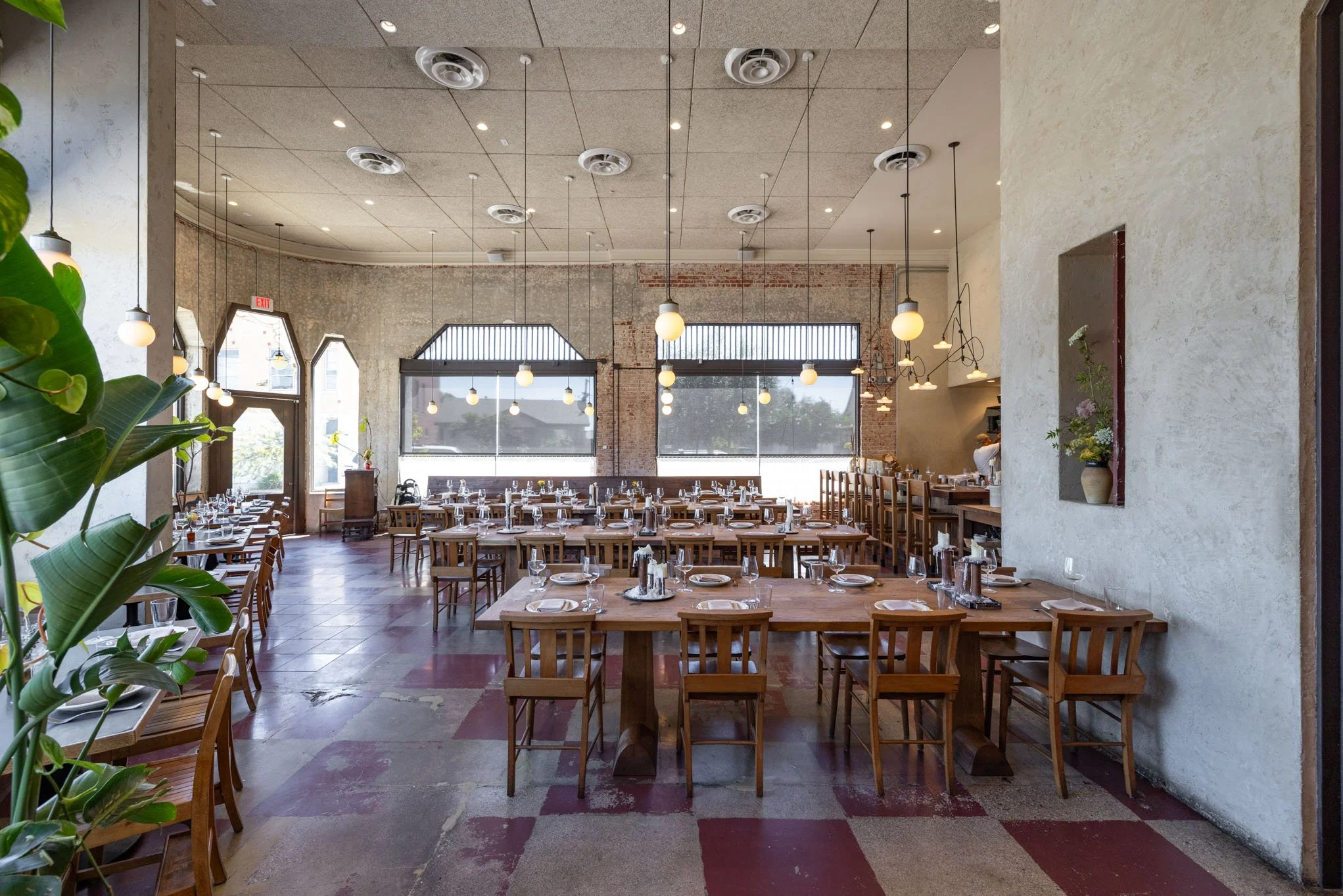 Interior of a spacious restaurant with wooden tables and chairs, set with glassware and plates, large windows, exposed brick walls, hanging pendant lights, and a decorative niche with a flower arrangement.