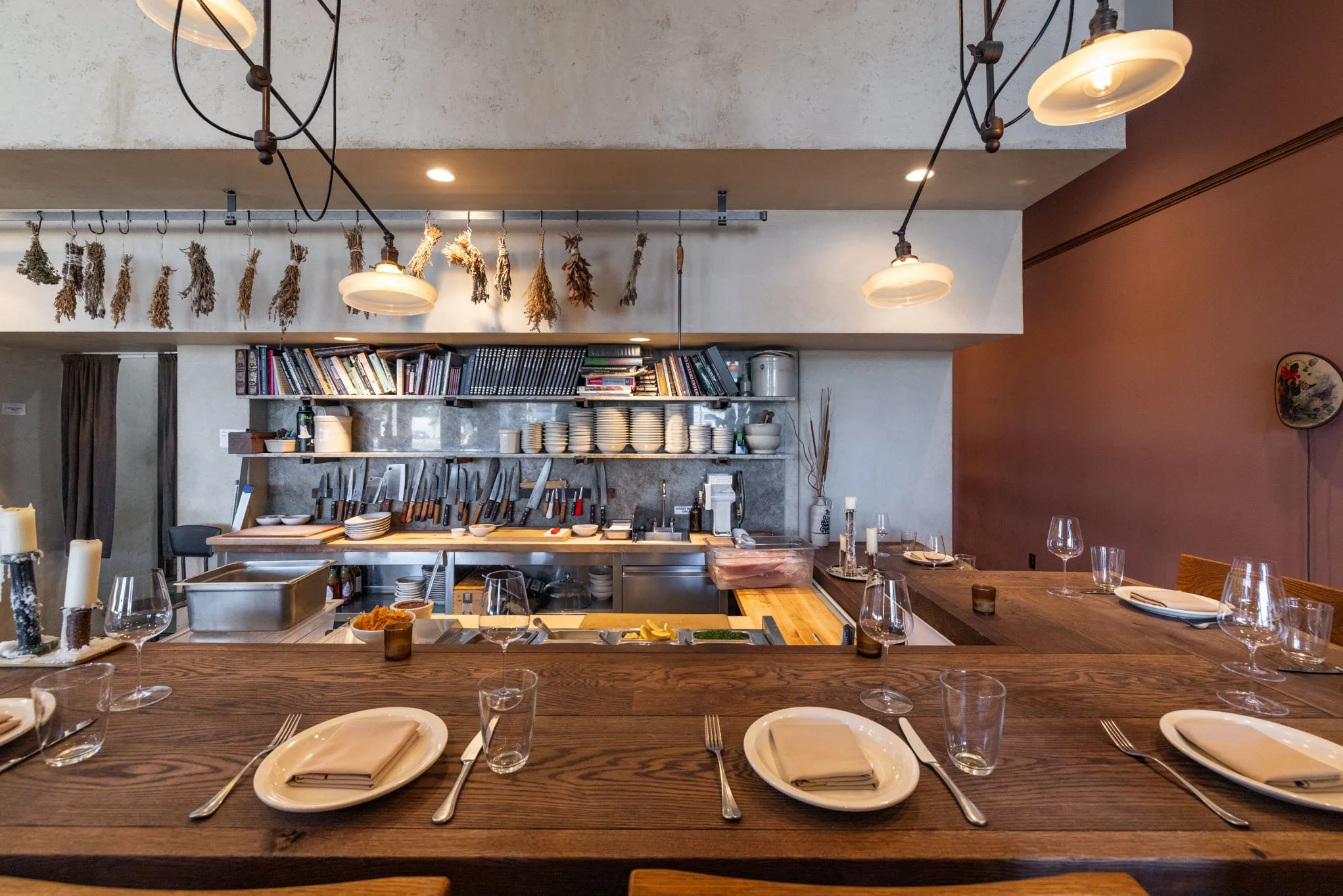 Empty dining table with plates, glasses, and silverware in a restaurant kitchen with hanging lights, shelves, and utensils.