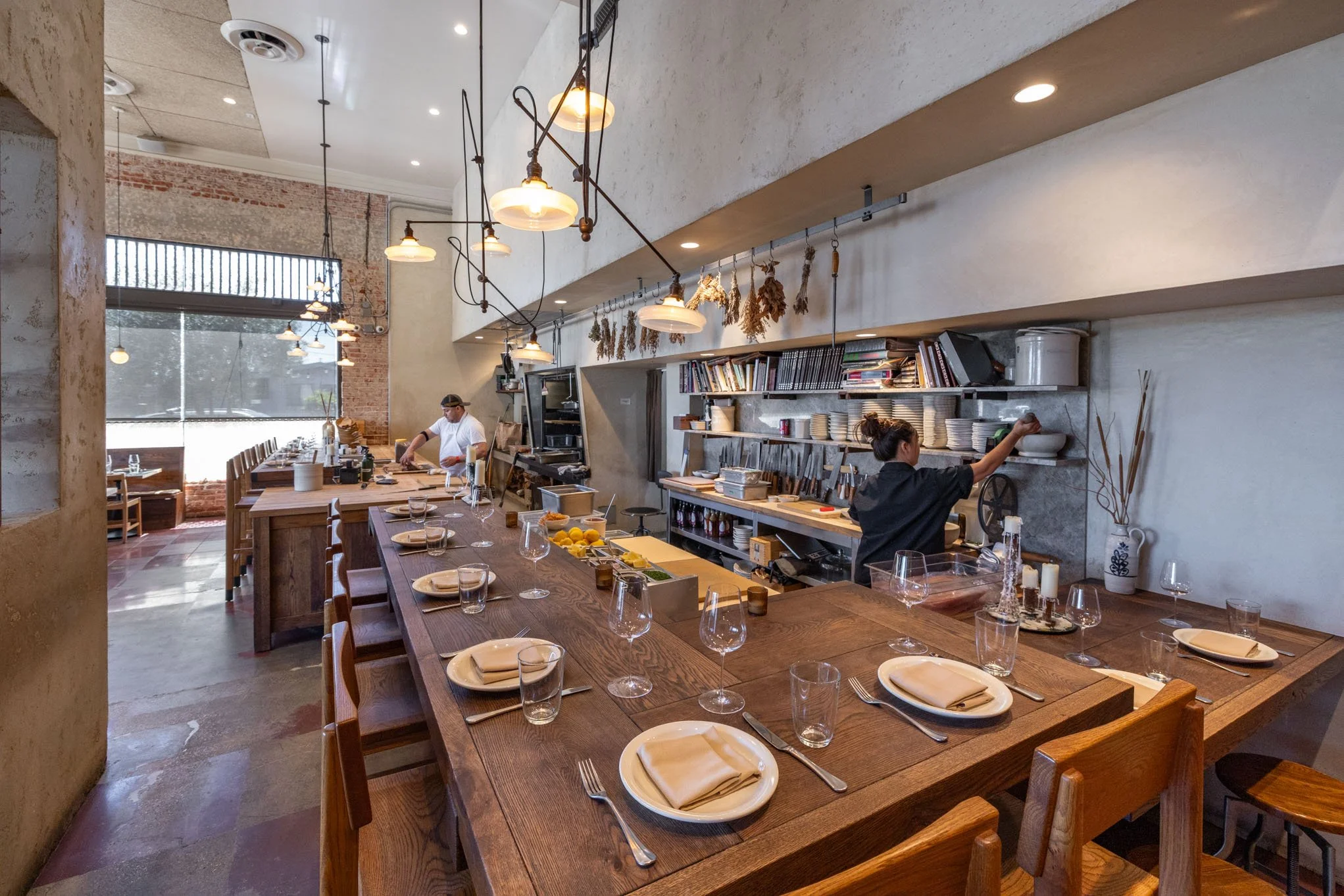 Interior of a modern, rustic restaurant with a large wooden communal table set with plates, napkins, and glasses, with a kitchen area in the background where a chef and a staff member prepare food.