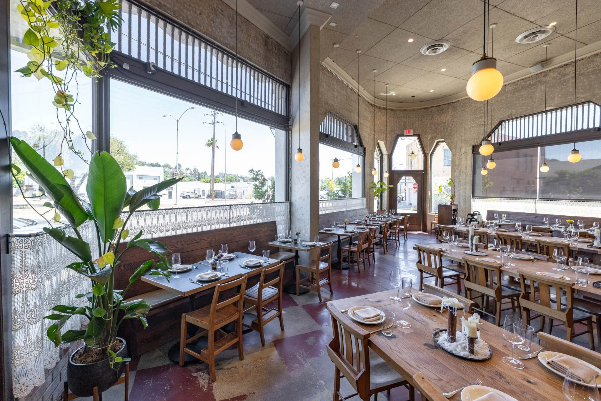 Dining area inside a restaurant with wooden tables and chairs, large windows, hanging pendant lights, potted plants, and table settings with plates, forks, knives, and wine glasses.