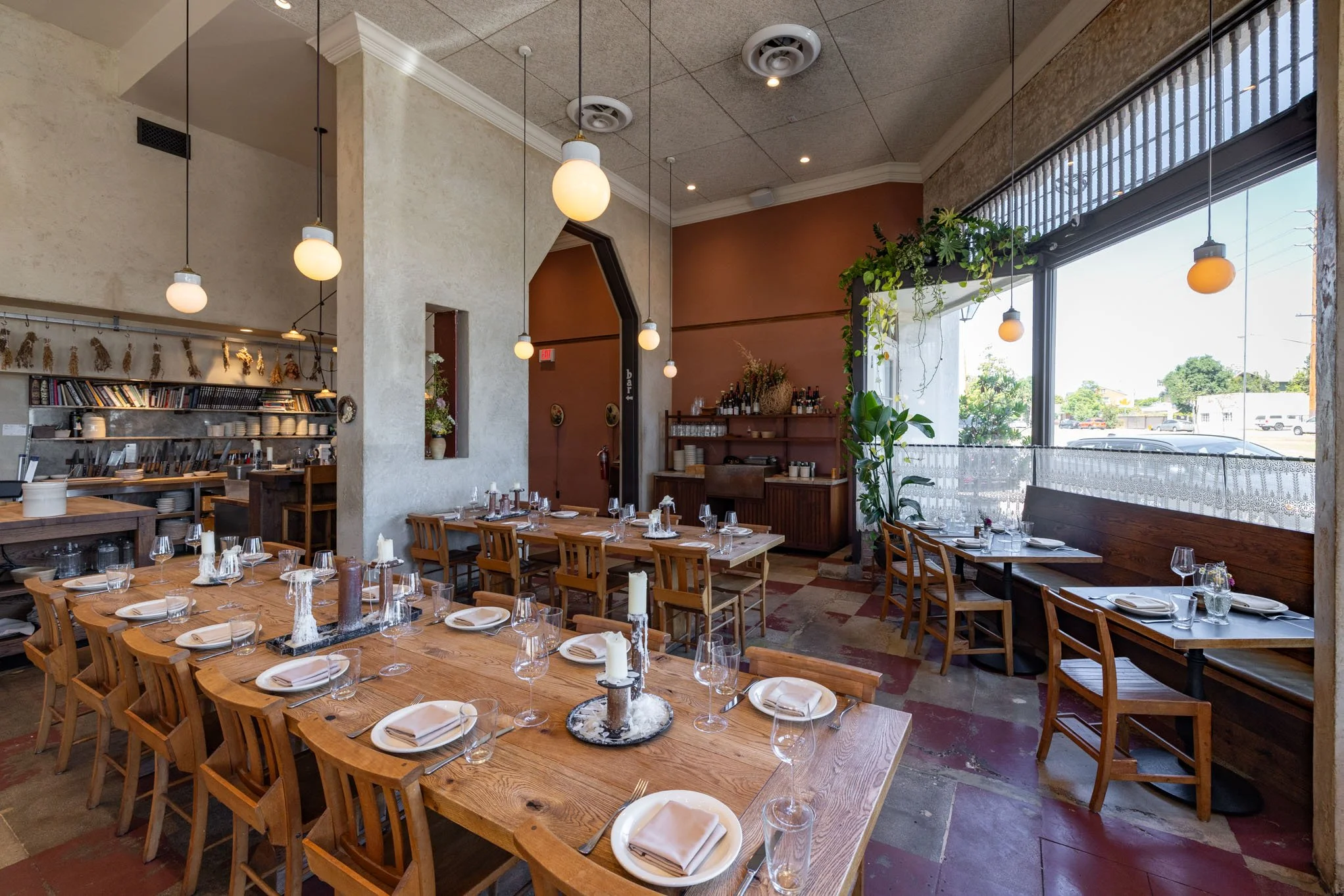 Interior of a restaurant with wooden tables set with plates, glasses, napkins, and candles. Large window with overhead lighting, potted plants, and a wall with shelves of books and decor.