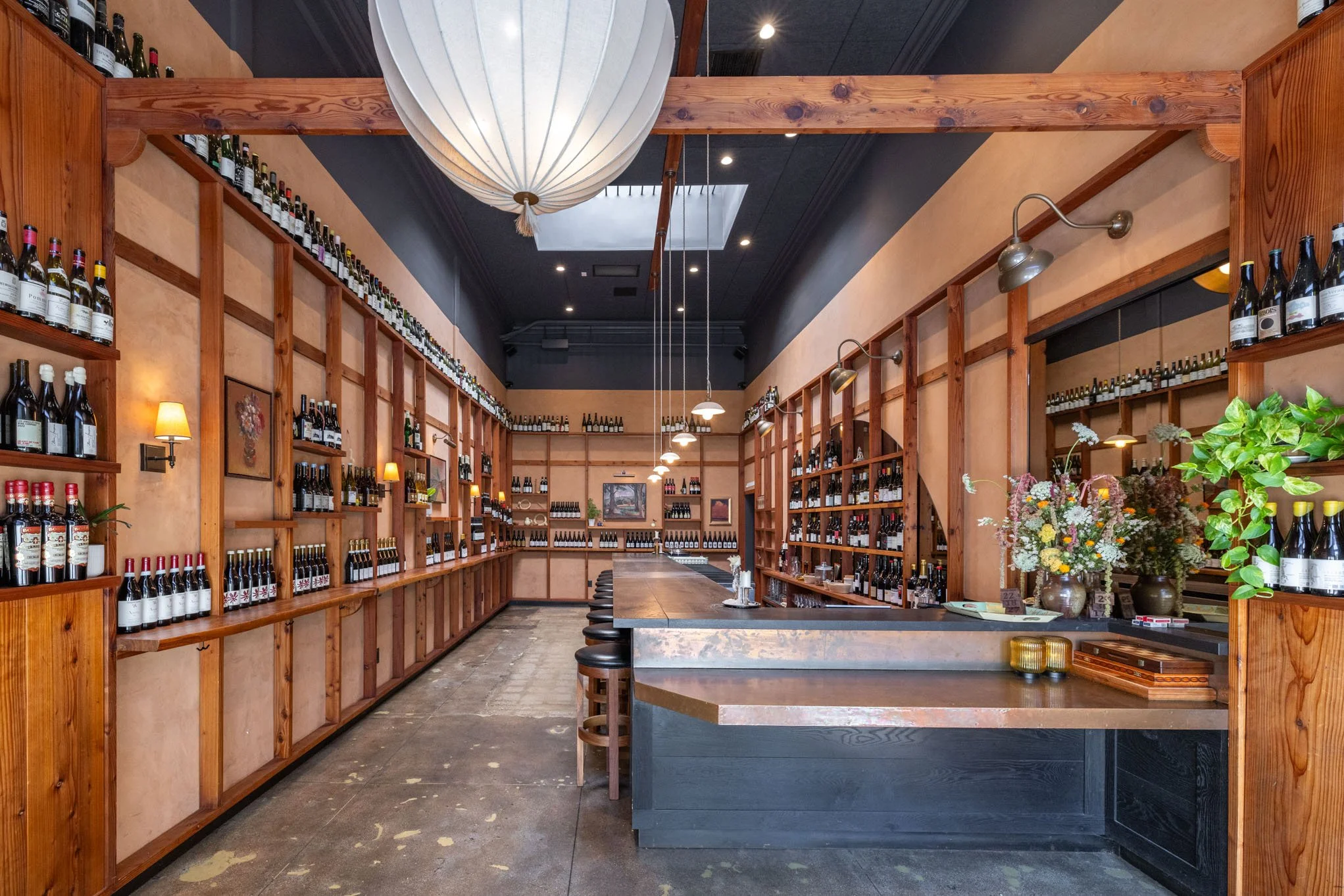 Interior of a wine shop with wooden shelves filled with bottles of wine, a long bar counter with stools, and decorative flowers, illuminated by pendant lights.