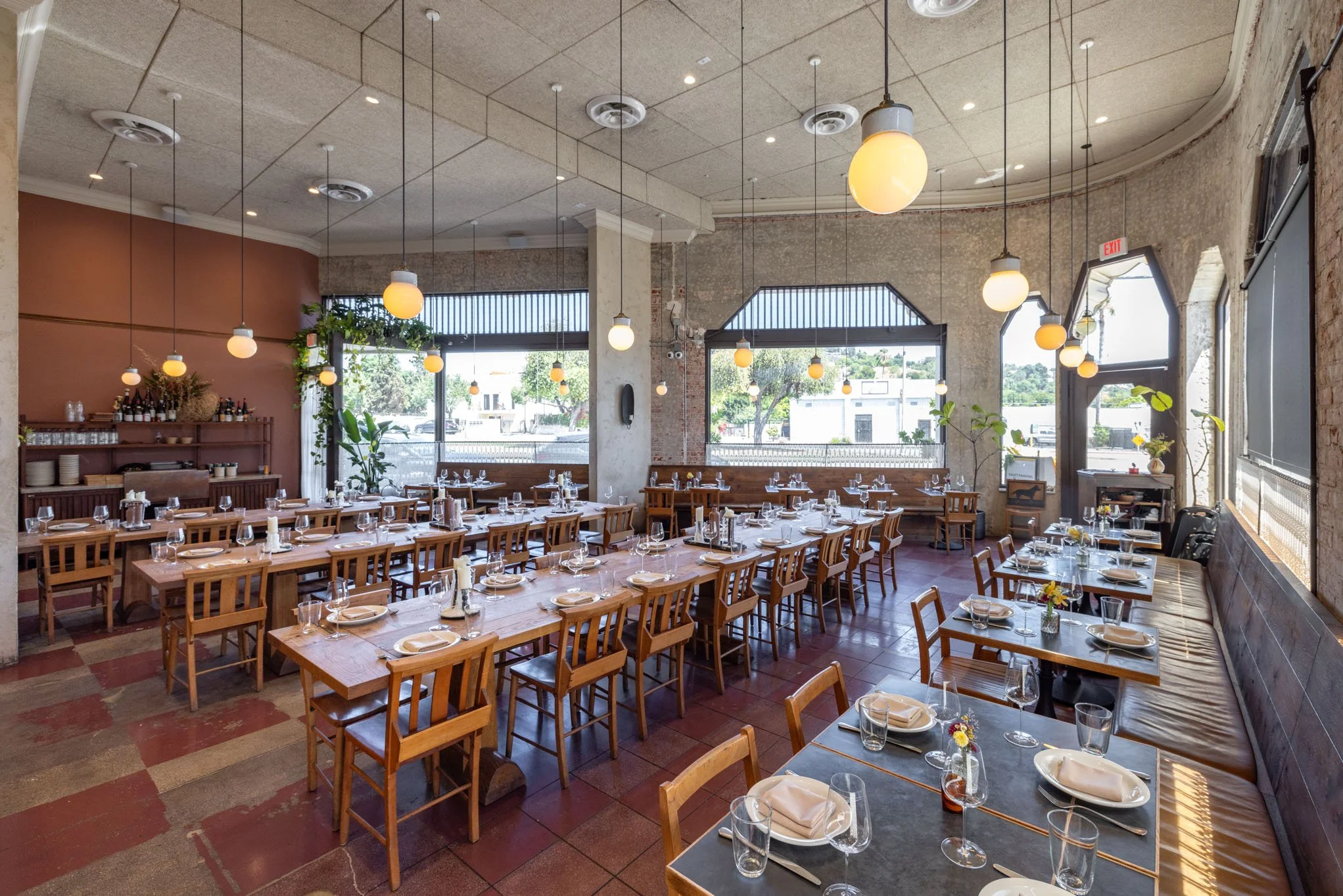 Interior of a modern restaurant with wooden tables, chairs, and place settings, natural light from large windows, hanging spherical pendant lights, potted plants, and a rustic accent wall.