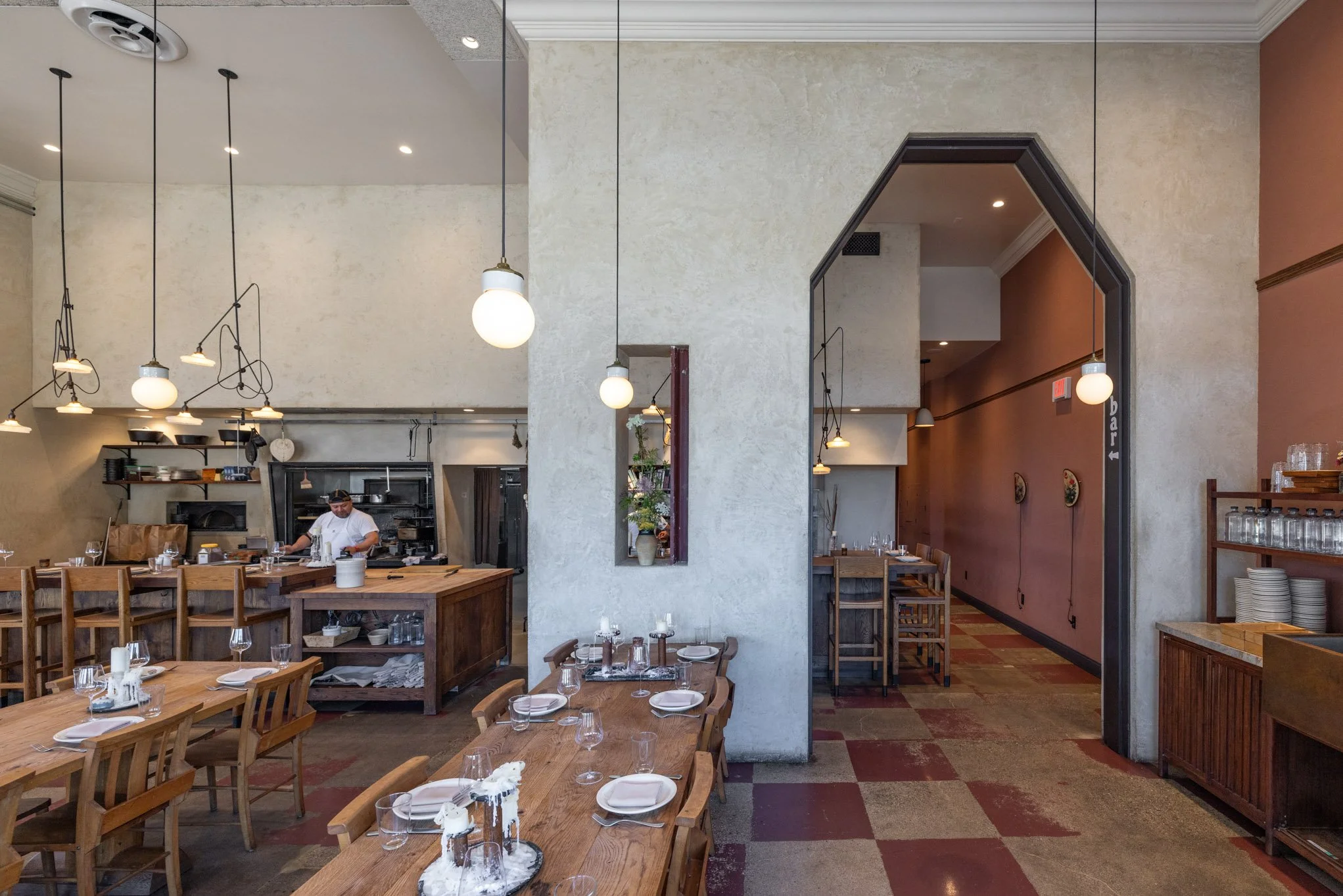 Interior of a restaurant with wooden tables set for dining, a visible kitchen in the background with a chef, and decorative hanging lights.