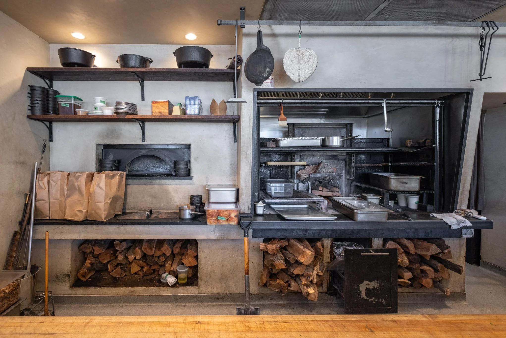A rustic kitchen with open shelves holding bowls, plates, and kitchen utensils above a wood-fired oven, with firewood stacked underneath and cooking pans hanging above.