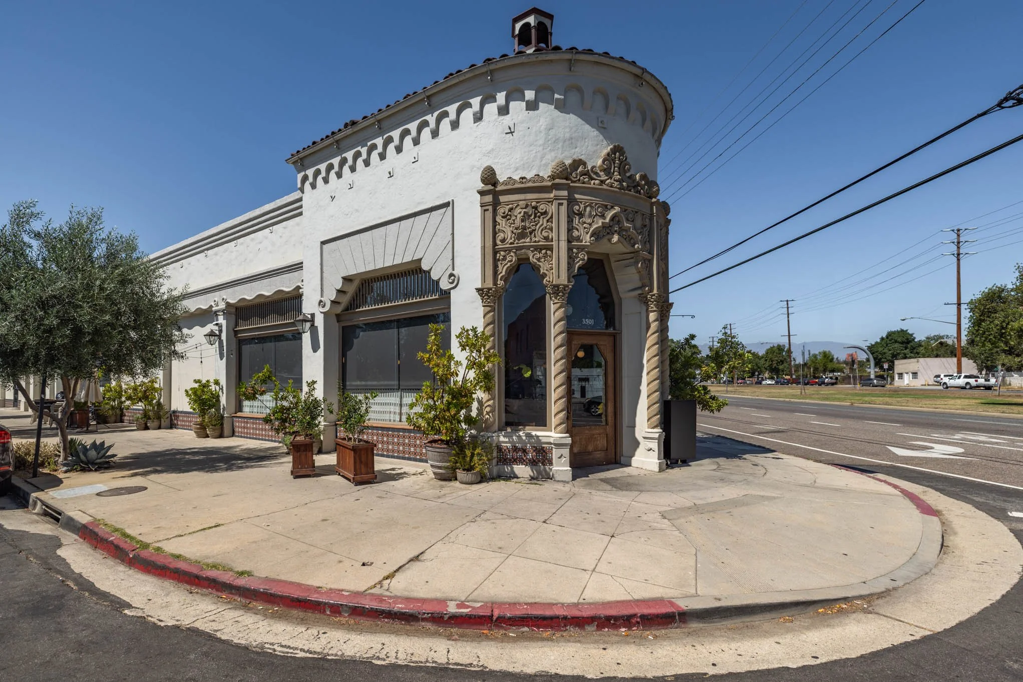 White building with ornate architectural details on the corner of a street, surrounded by potted plants and trees, with a clear blue sky in the background.