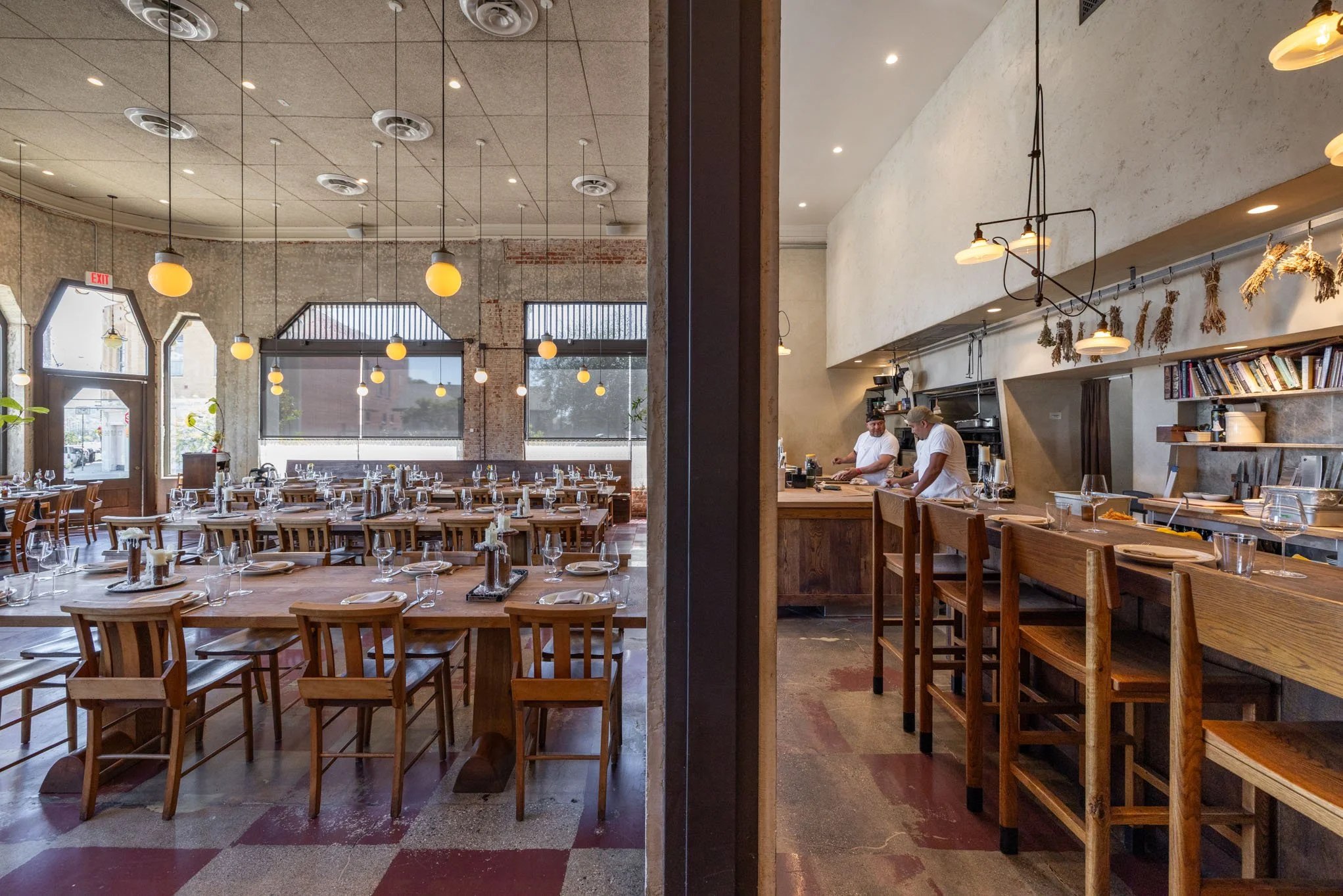 Interior of a restaurant with tables set for dining and two chefs preparing food in the kitchen area.